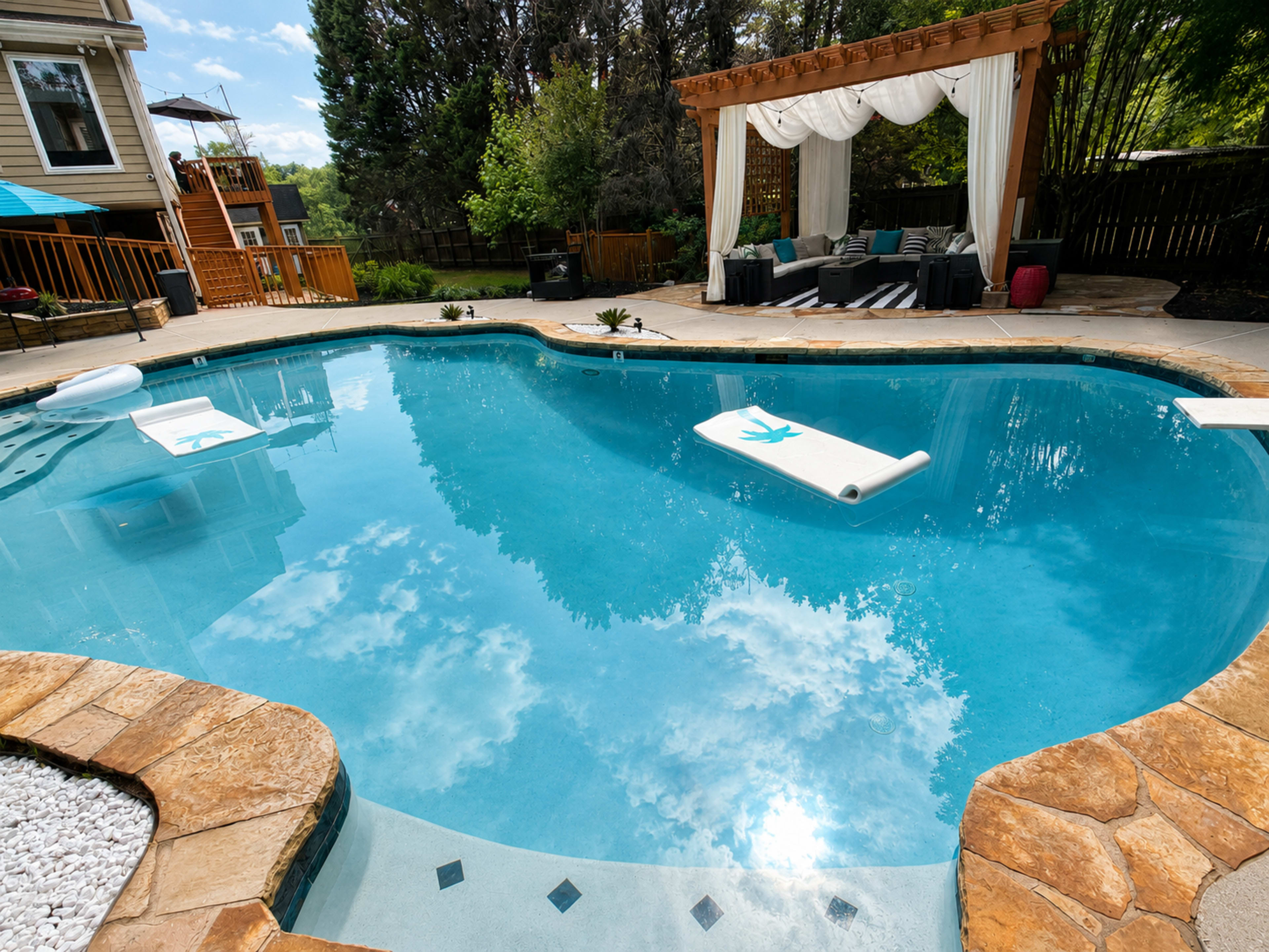 The image shows a well-designed backyard pool area with a stone patio, lounge floats in the water, and a shaded seating area under a wooden pergola.