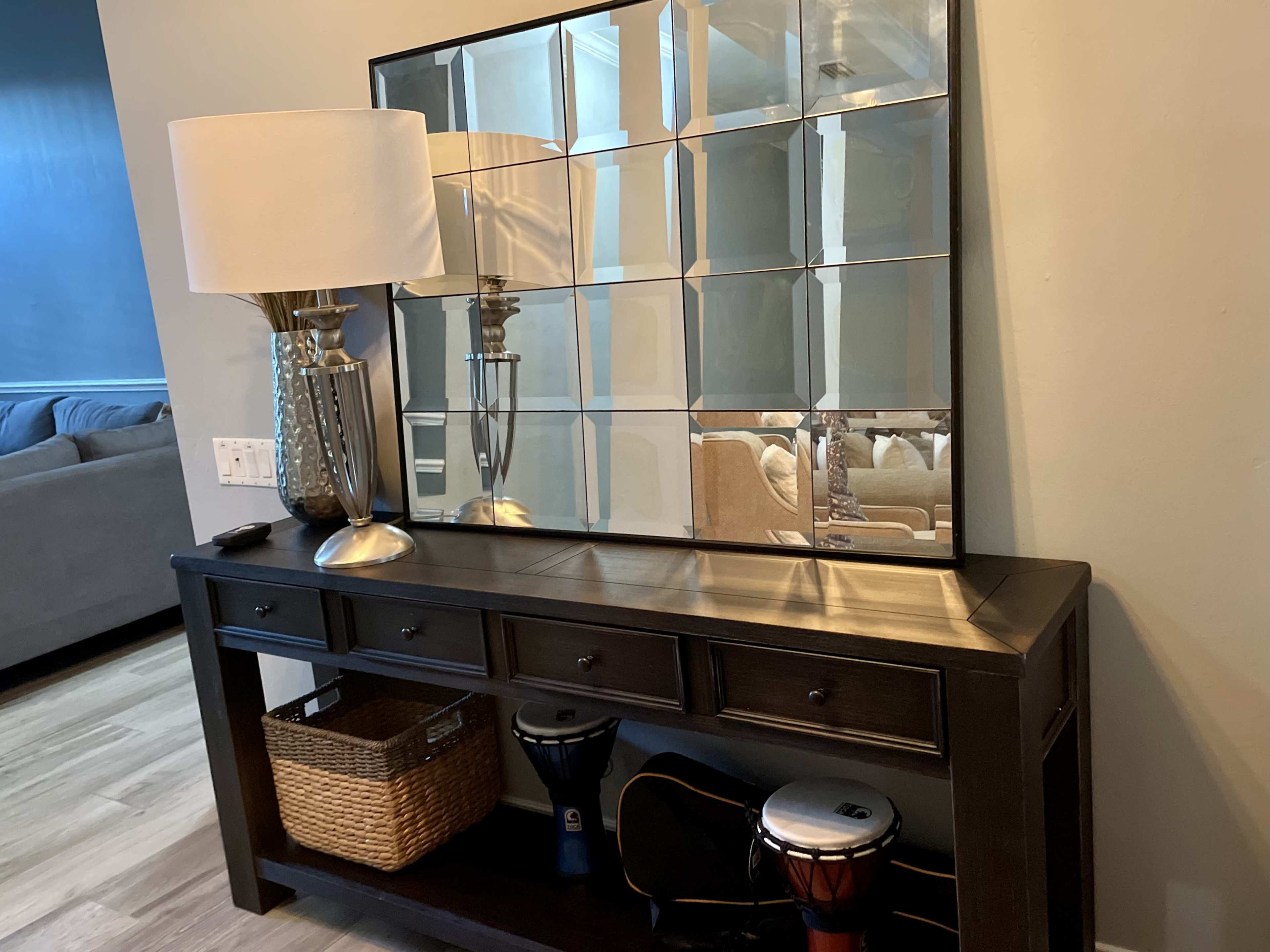 A dark wooden console table with drawers, a large mirror above it, a decorative lamp, and a wicker basket beside a pair of drums.