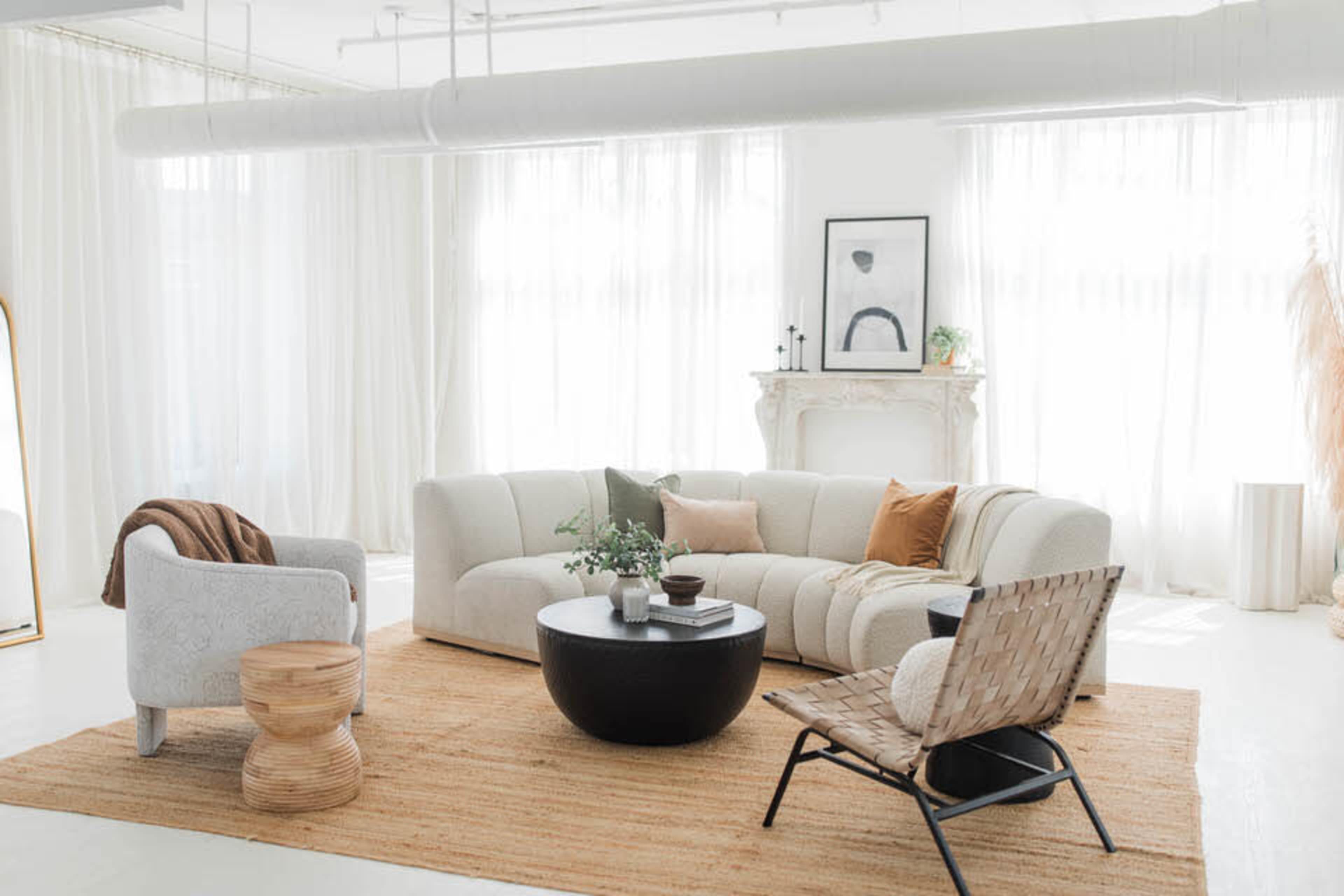 The image shows a bright and airy living room with a curved sofa, a round coffee table, and various seating options on a jute rug, accented by large windows and decorative elements.