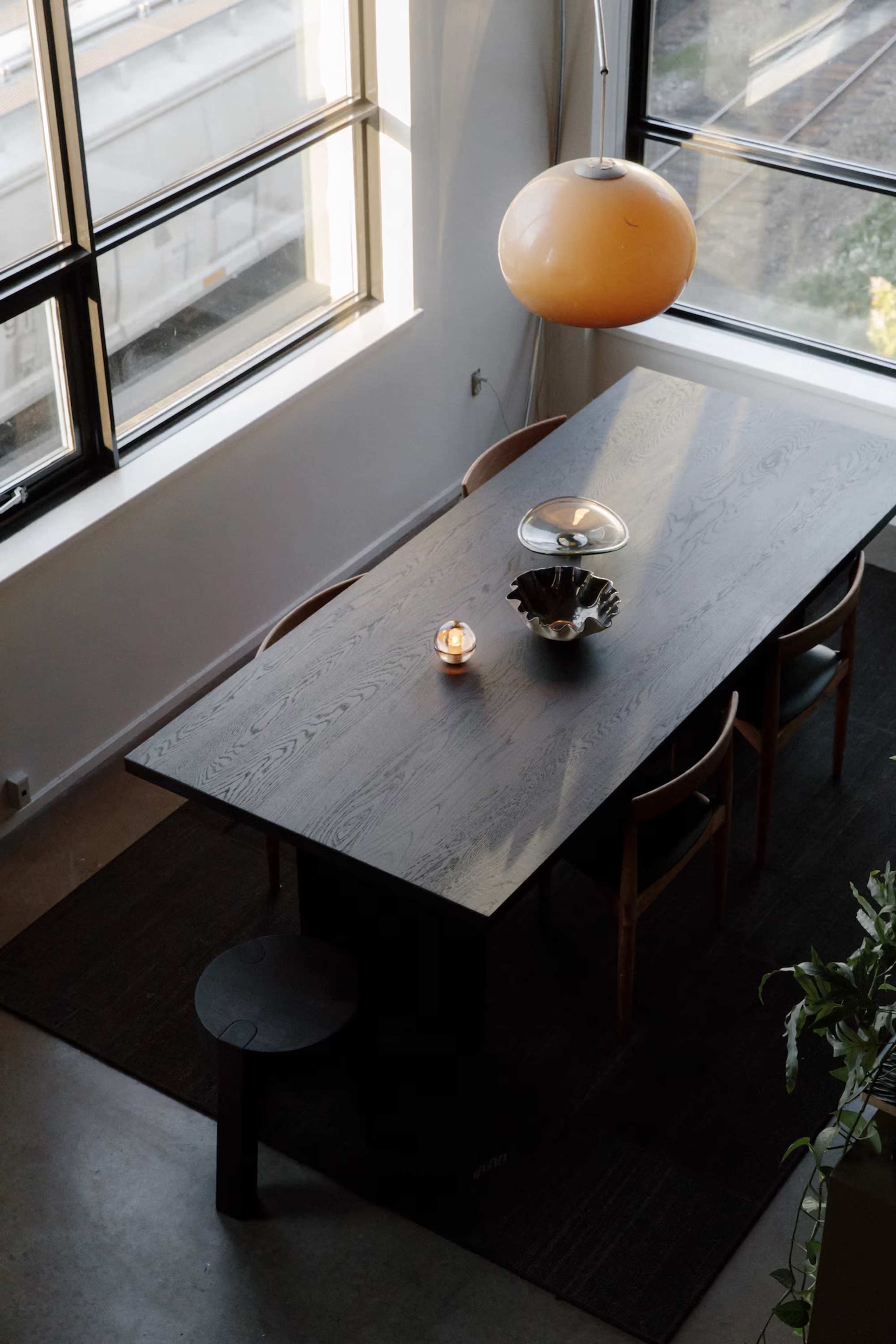 A wooden dining table with chairs is positioned under a pendant light in a well-lit room featuring large windows.