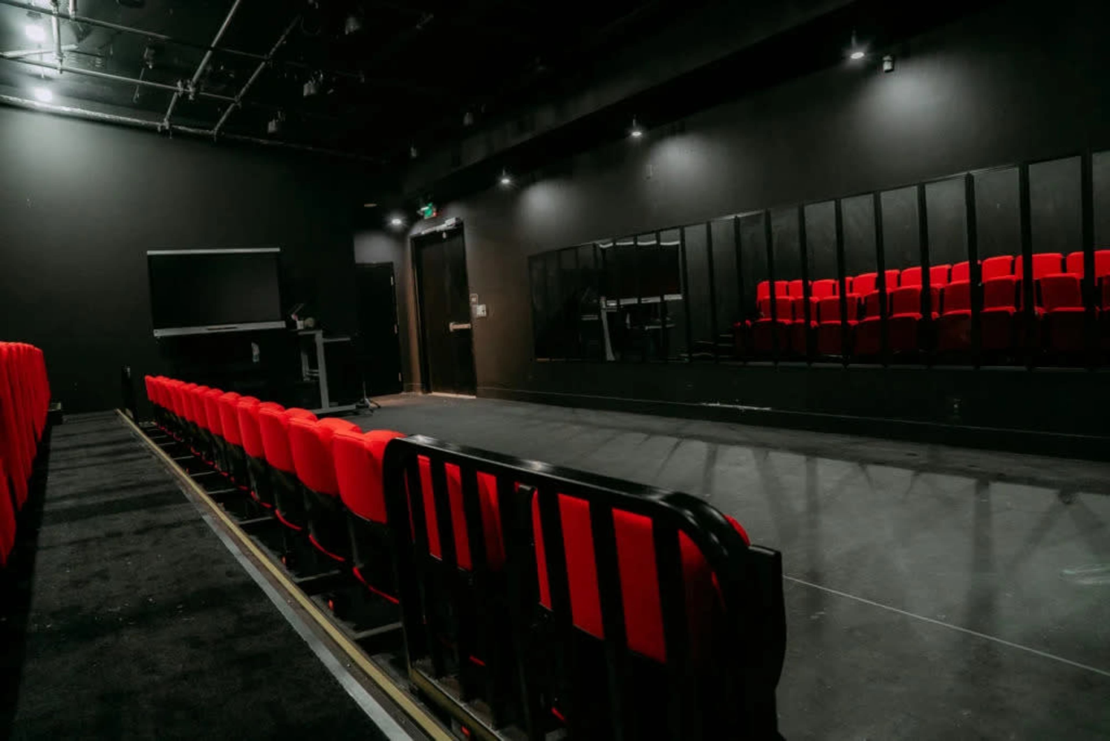 The image depicts a small black-walled theater with red seats arranged in rows facing a performance area and a large mirror on one side.
