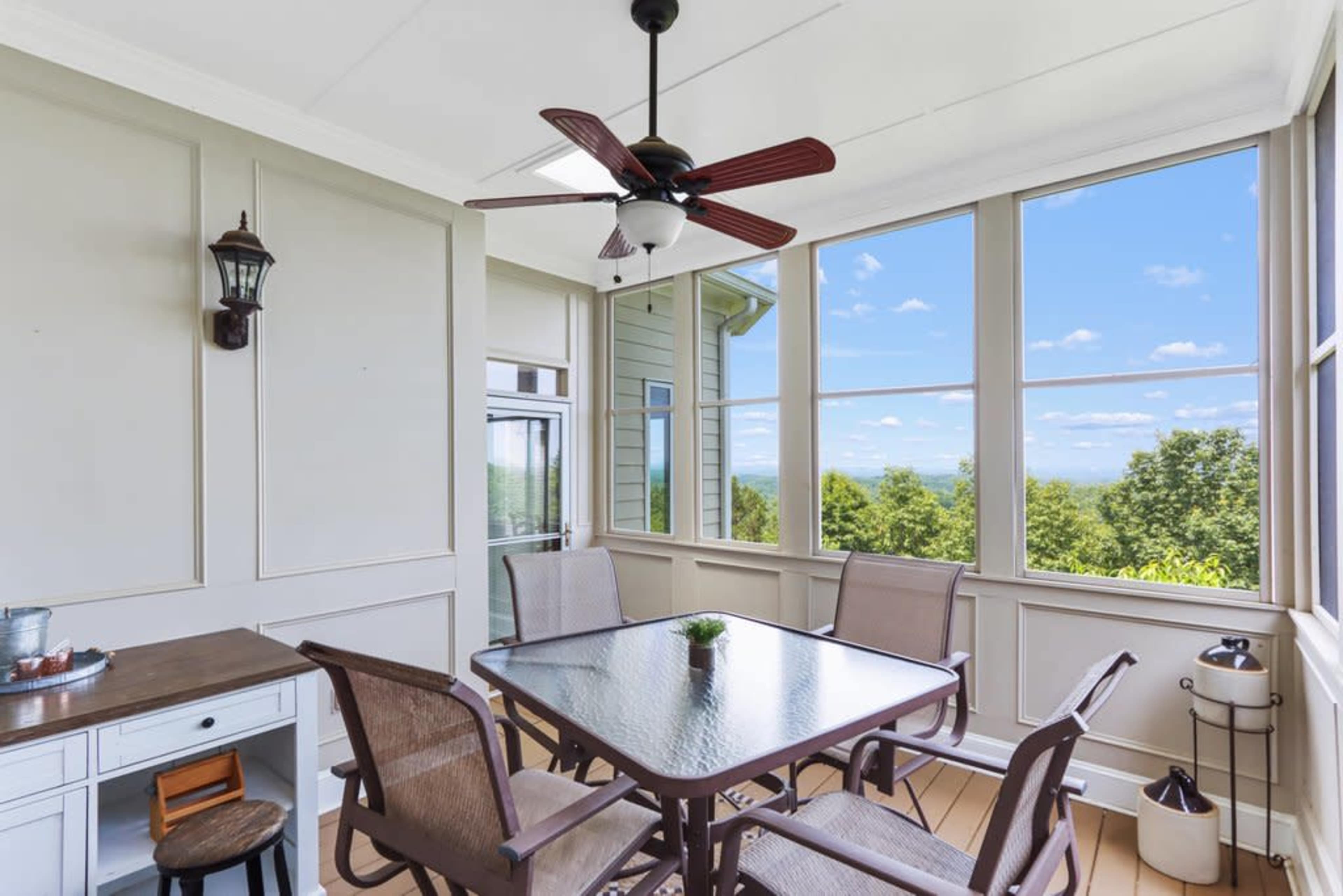 A sunlit sunroom with large windows, a ceiling fan, a glass table, and several chairs, surrounded by greenery.