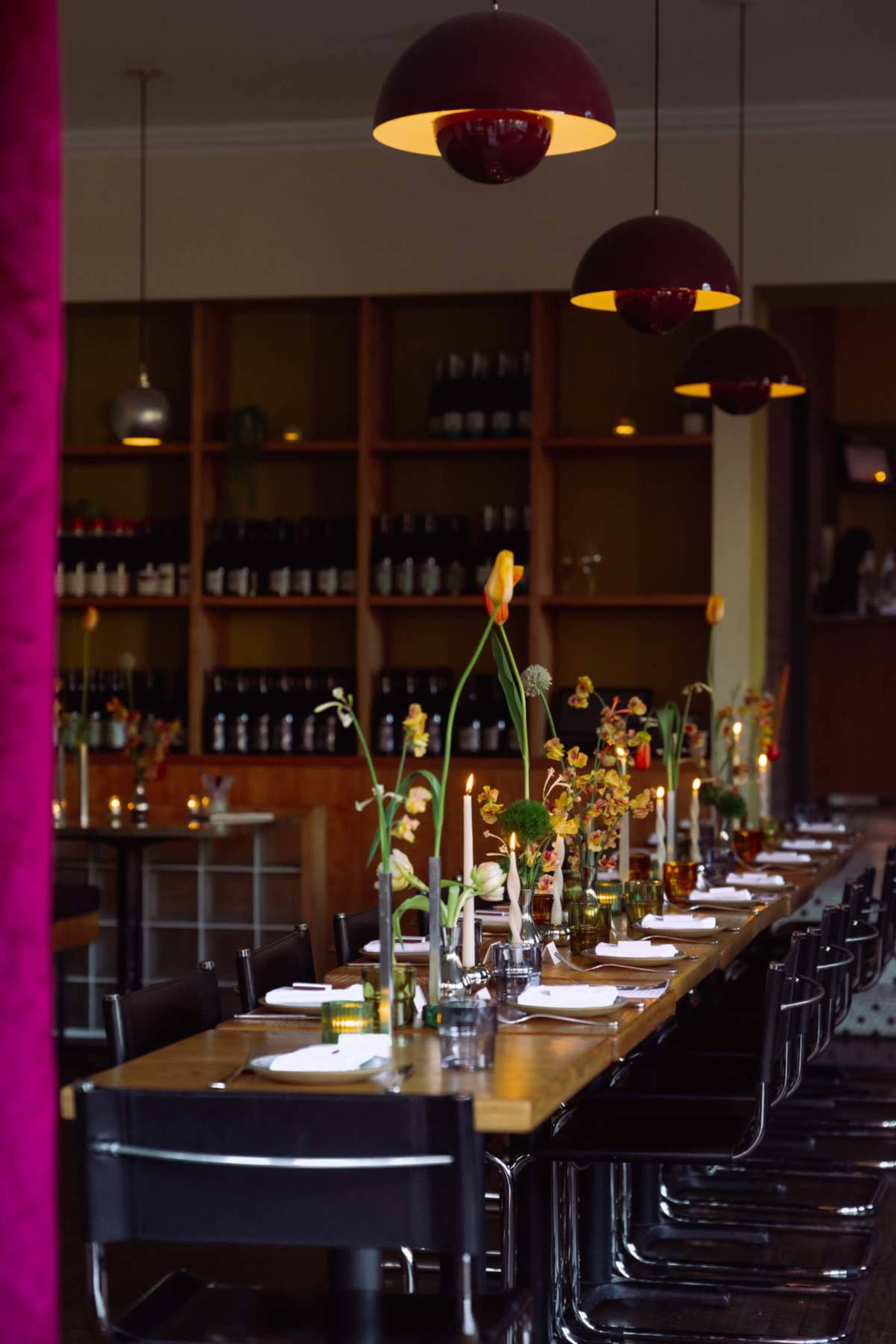 A long dining table is set with floral centerpieces and candles in a restaurant with shelves of wine in the background.