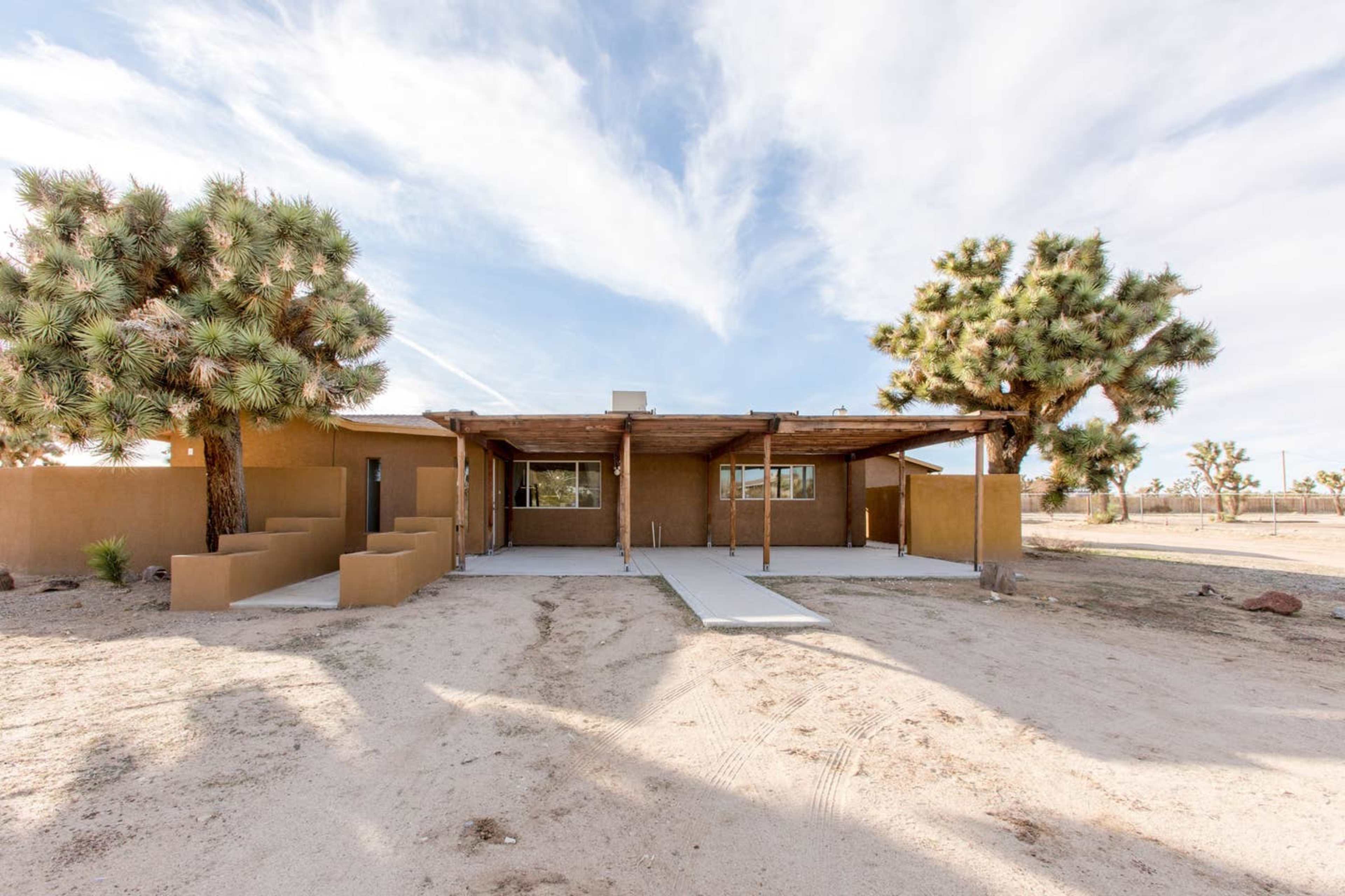 A single-story, adobe-style house with a wooden porch is set against a backdrop of Joshua trees and a clear sky.