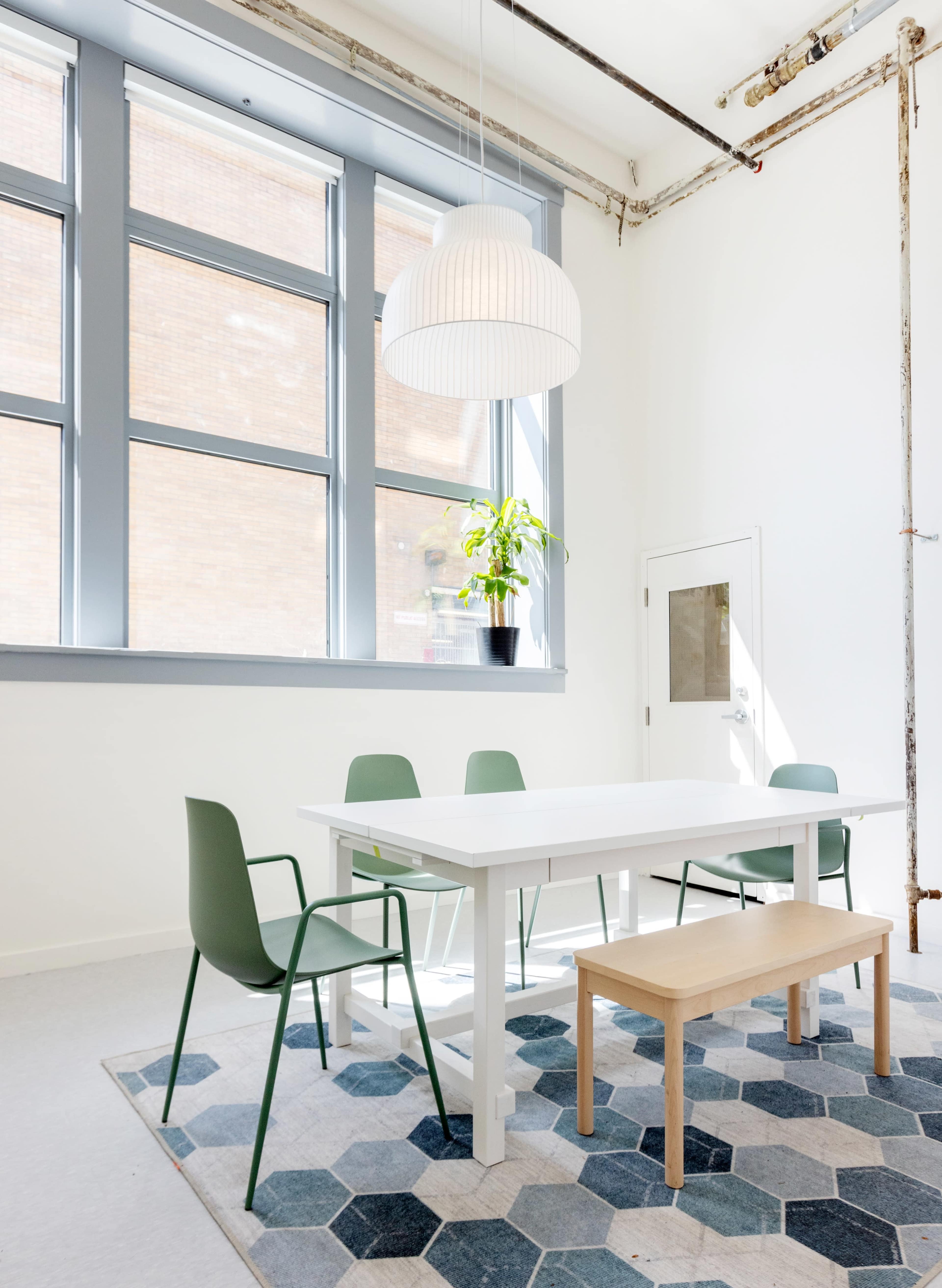 A modern dining area features a white table, green chairs, a light fixture, and a geometric rug under large windows.