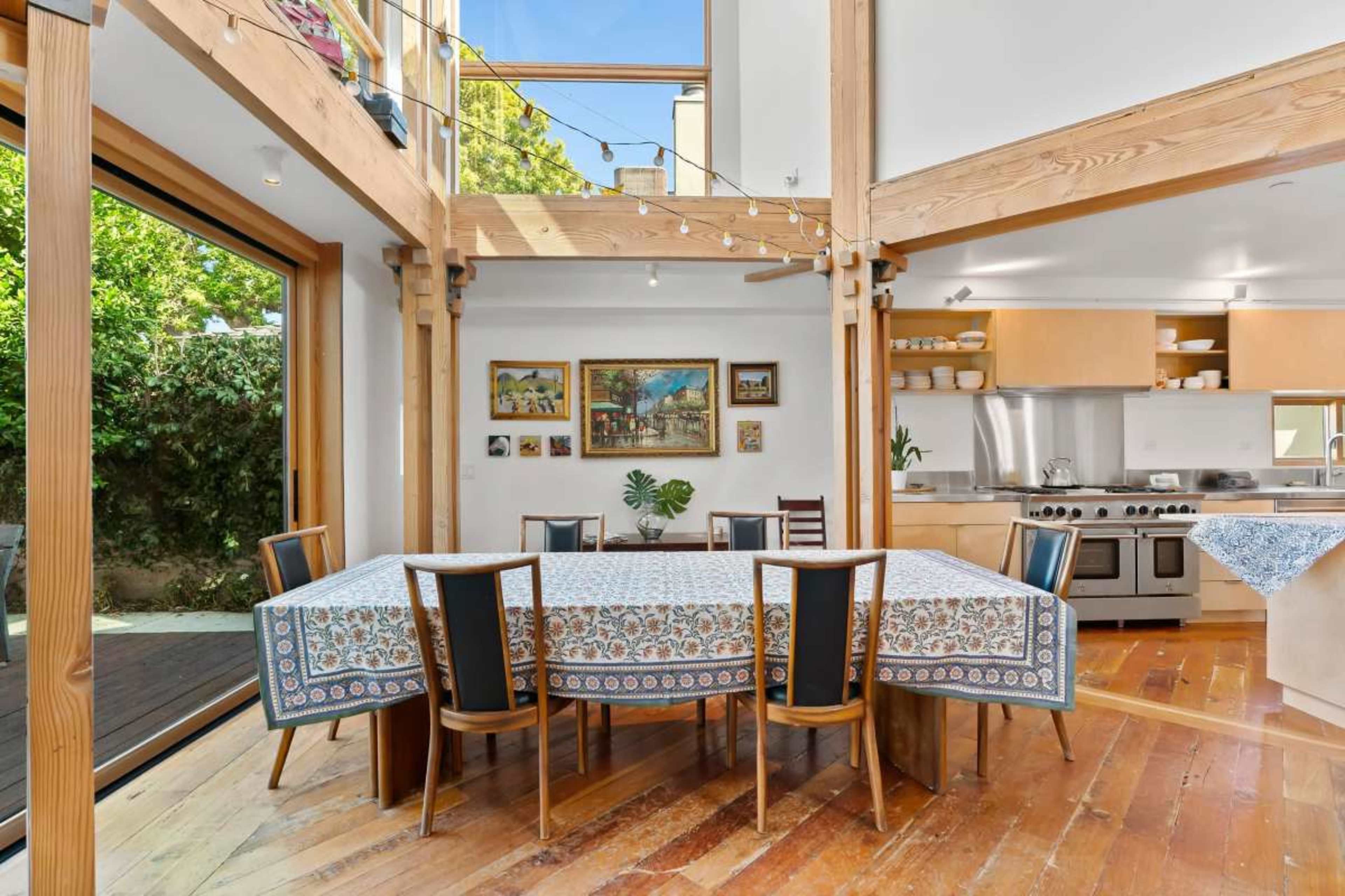 The image shows a spacious dining area with a large rectangular table set beneath wooden beams, adjacent to an open kitchen with modern appliances and a view of a landscaped garden through large glass doors.