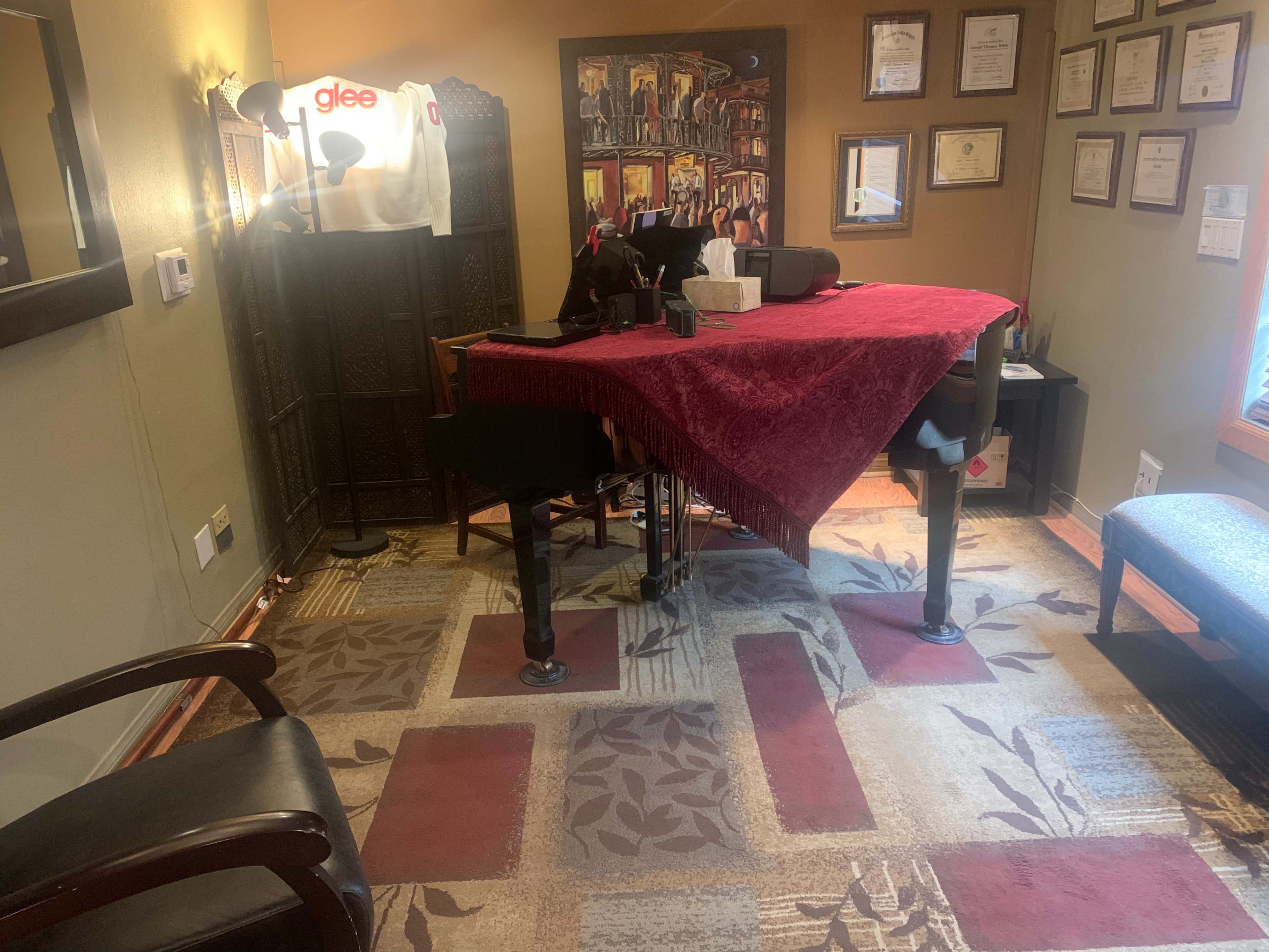 The image shows a room with a grand piano covered by a red cloth, a desk with various items, framed certificates on the walls, and a chair in the corner.