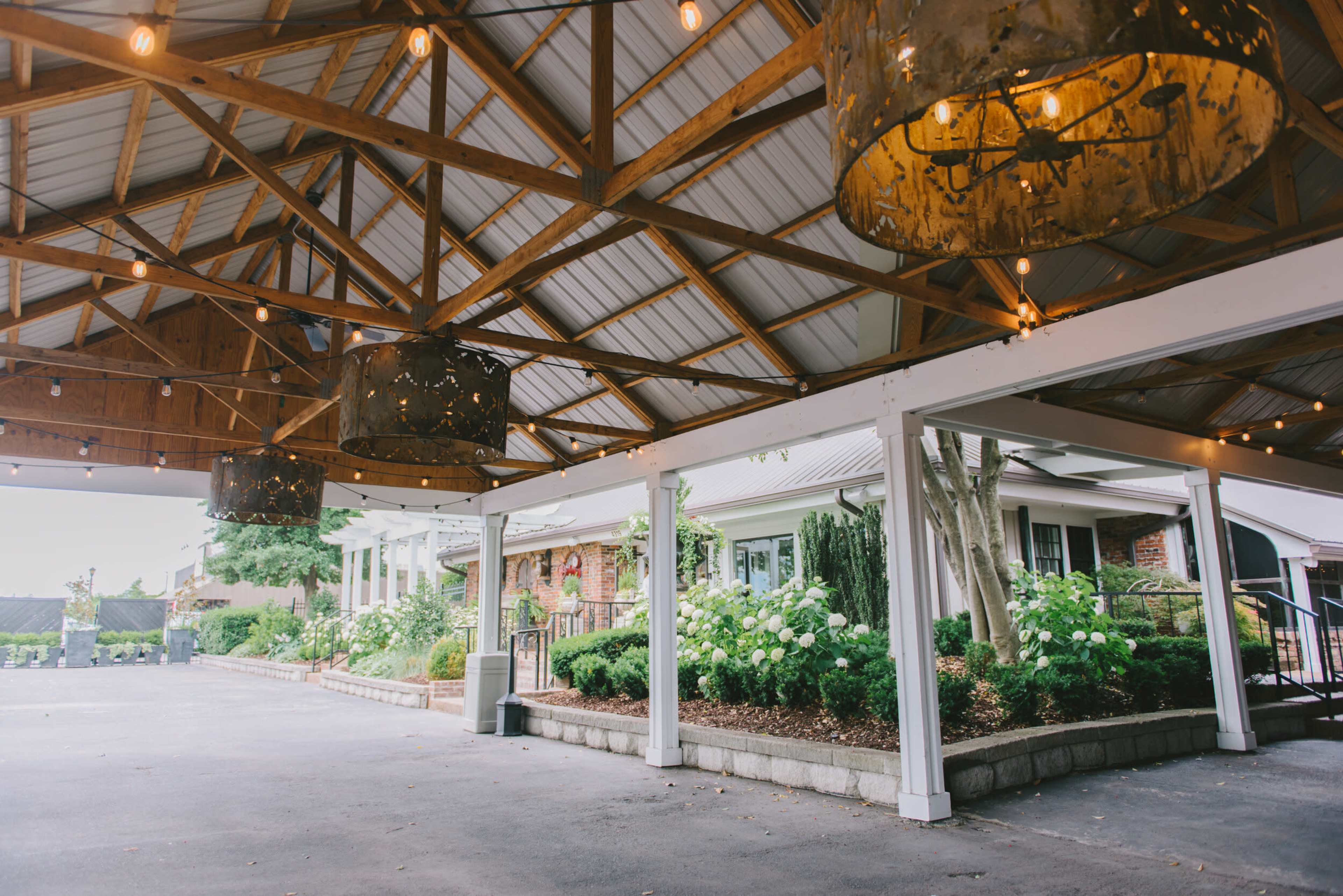 A covered outdoor space featuring wooden beams, hanging light fixtures, and a view of a landscaped area with greenery and flowers.