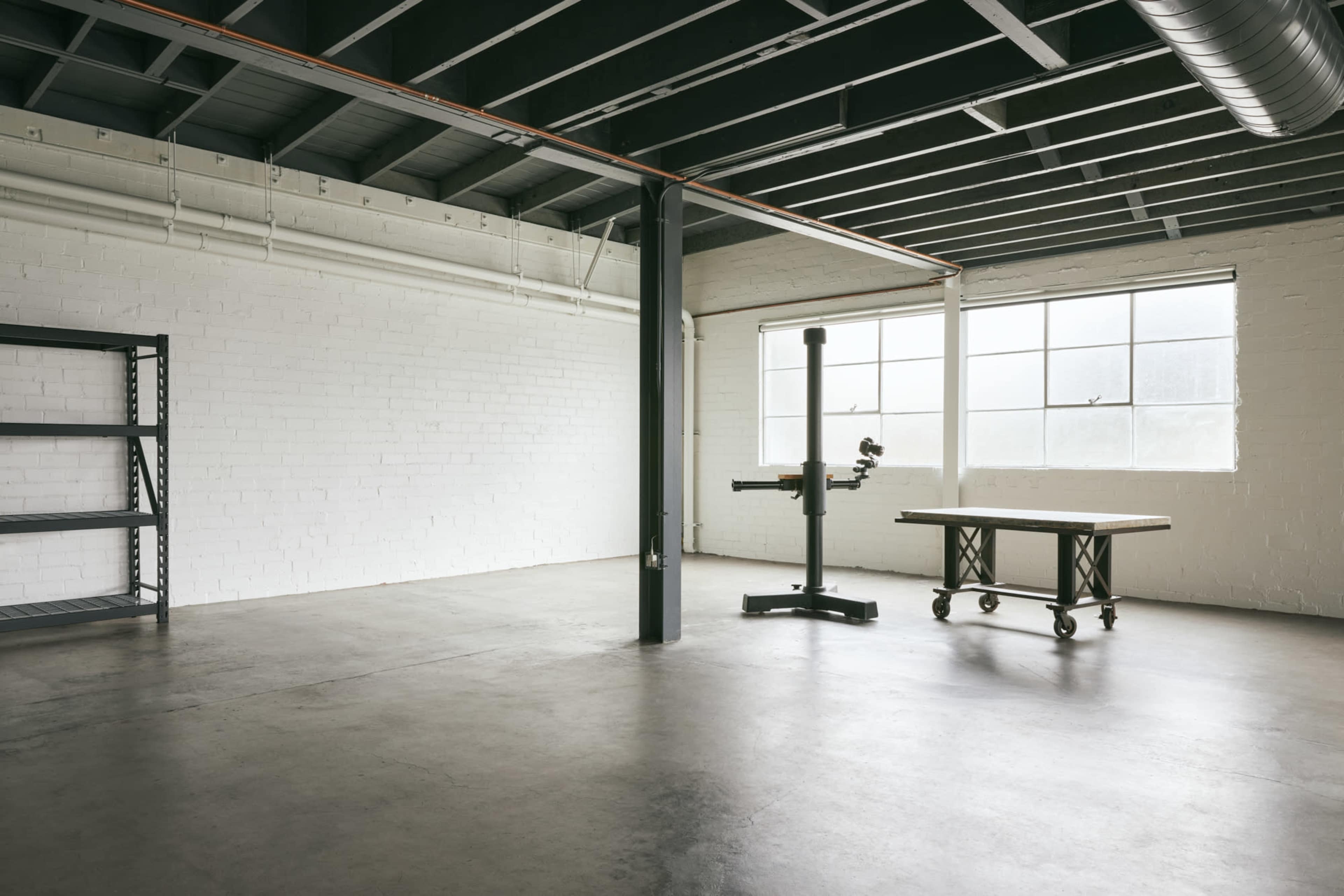 An empty industrial workspace features concrete flooring, exposed beams, a large window, a metal shelf, and a wooden table beside a machinery stand.