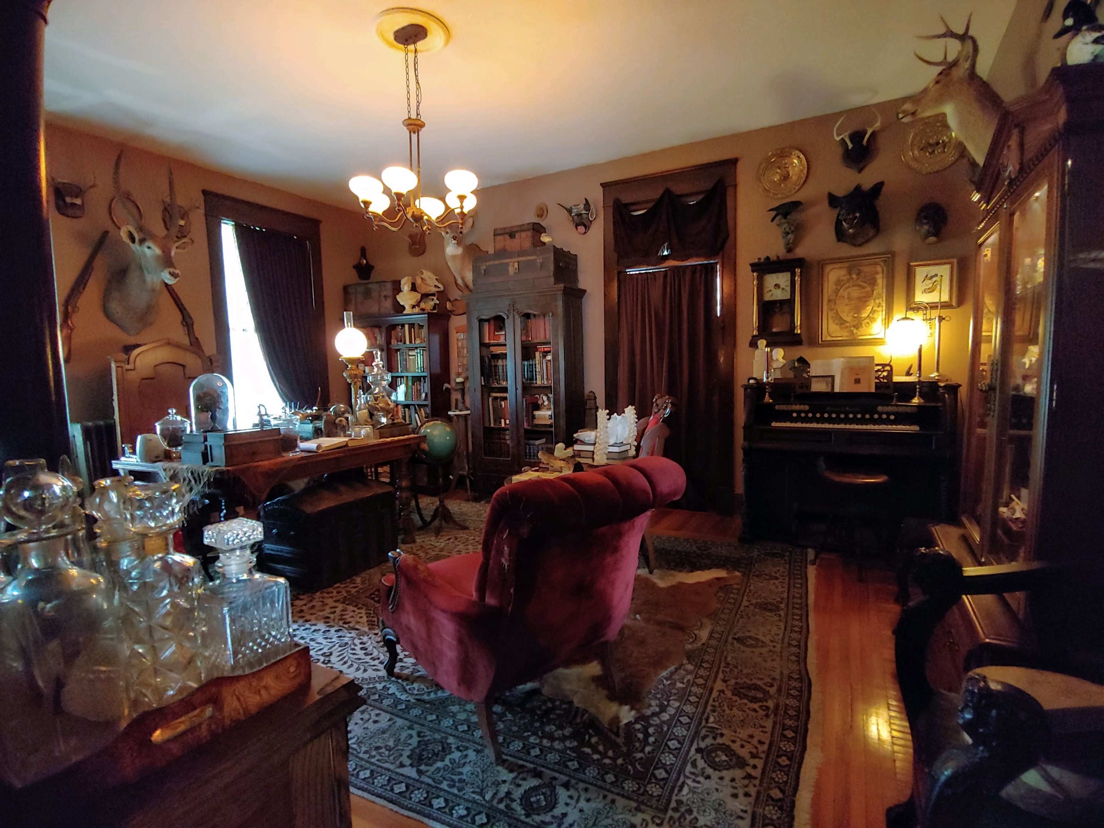 The room features vintage furniture, a bookshelf filled with books, and various mounted animal heads on the walls, illuminated by several lamps.