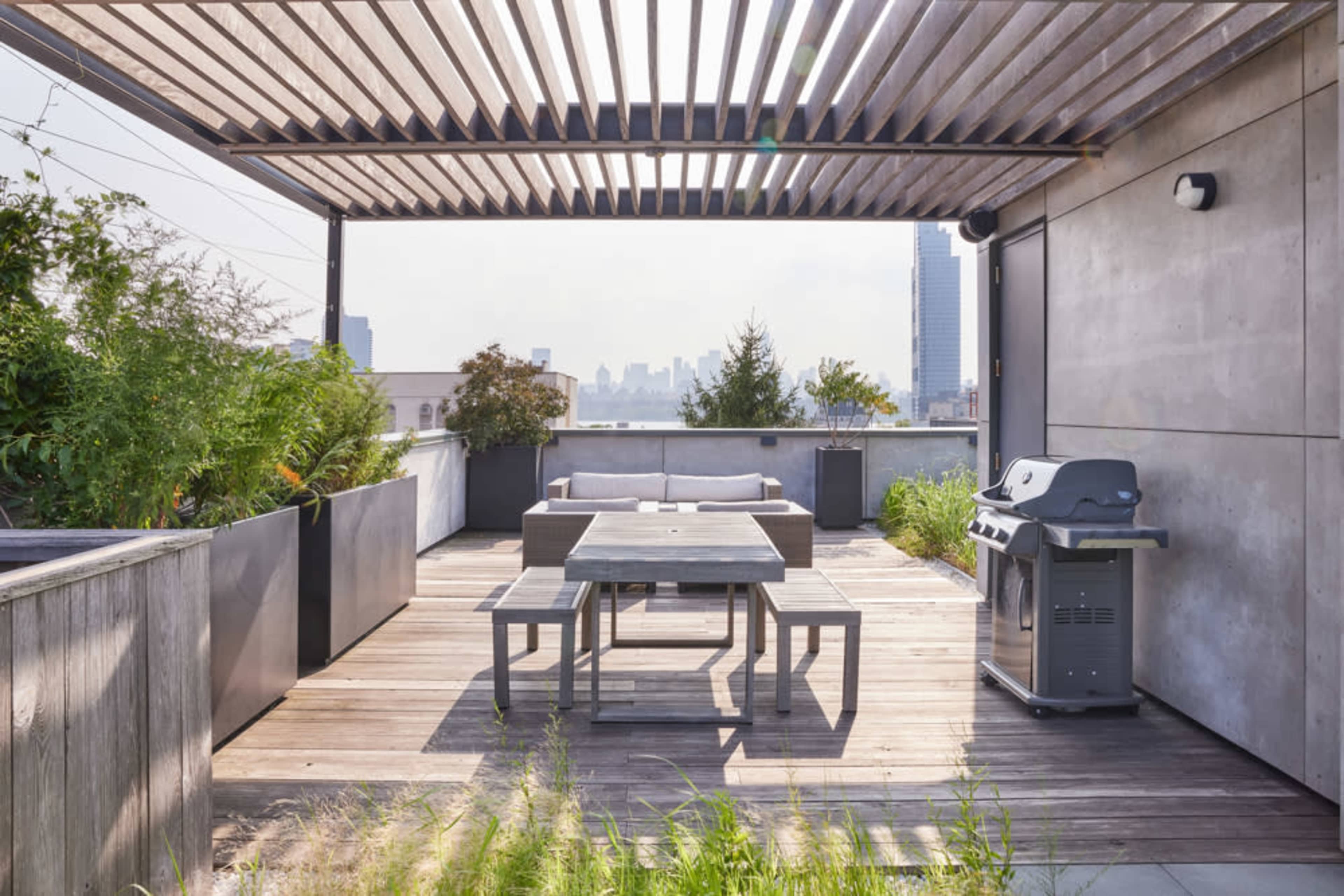 A rooftop terrace with wooden decking, a dining table, a barbecue grill, and planters filled with greenery, overlooking a city skyline.