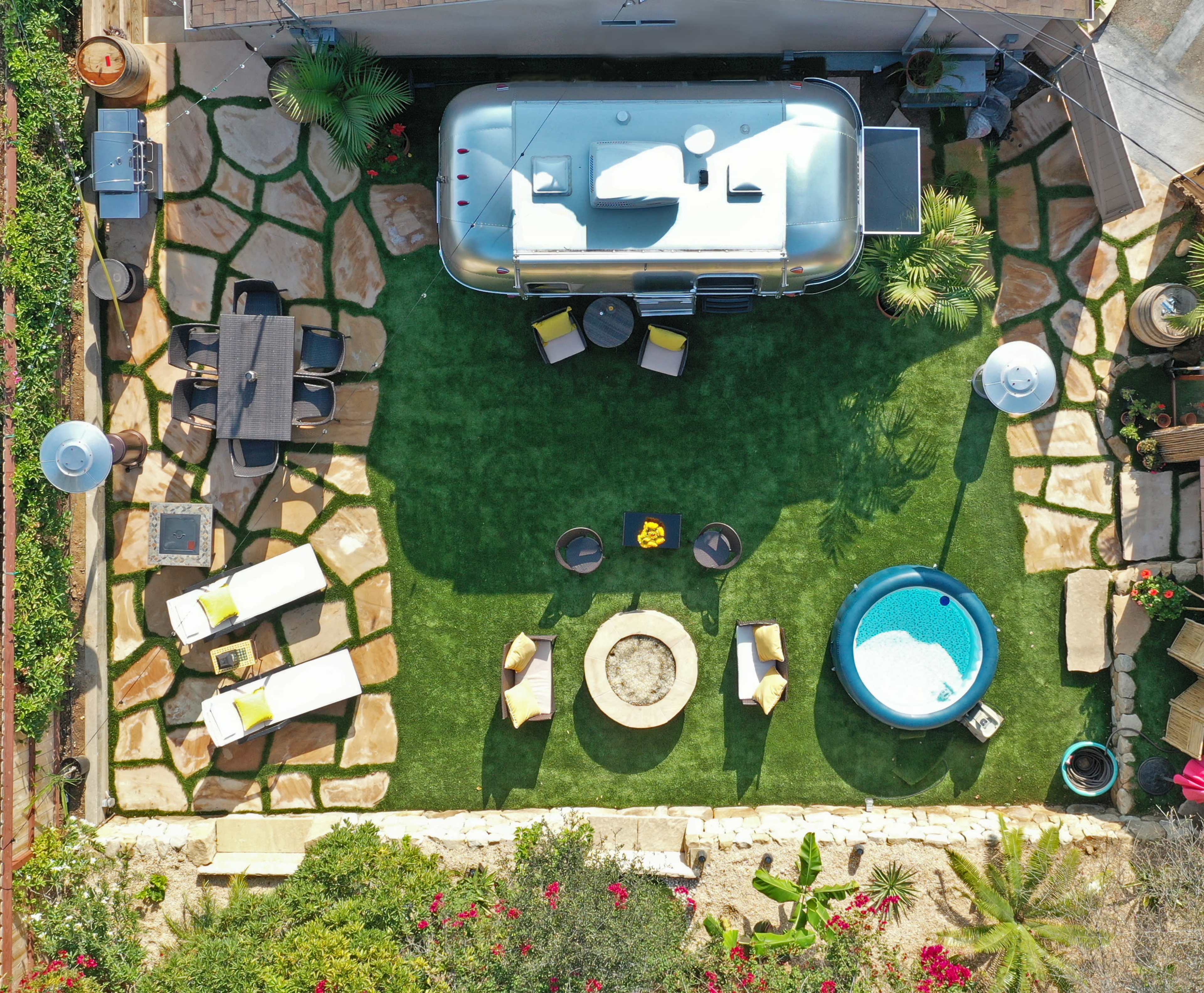 An outdoor patio area featuring a silver Airstream trailer, a circular hot tub, a fire pit, and various seating arrangements on stone and grassy surfaces.