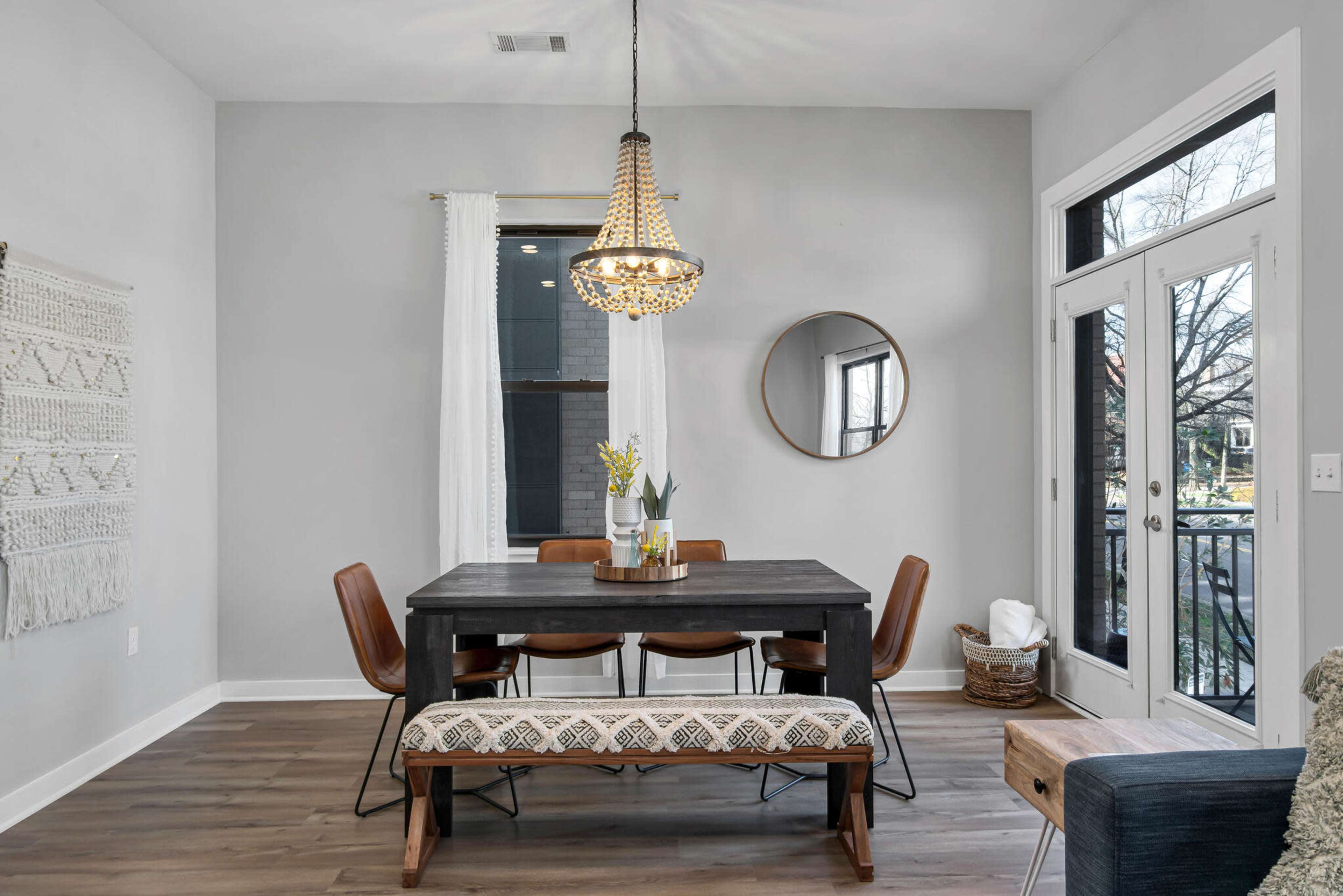The image shows a modern dining area featuring a wooden table with chairs and a bench, illuminated by a chandelier, with a large round mirror and macramé wall decor in the background.
