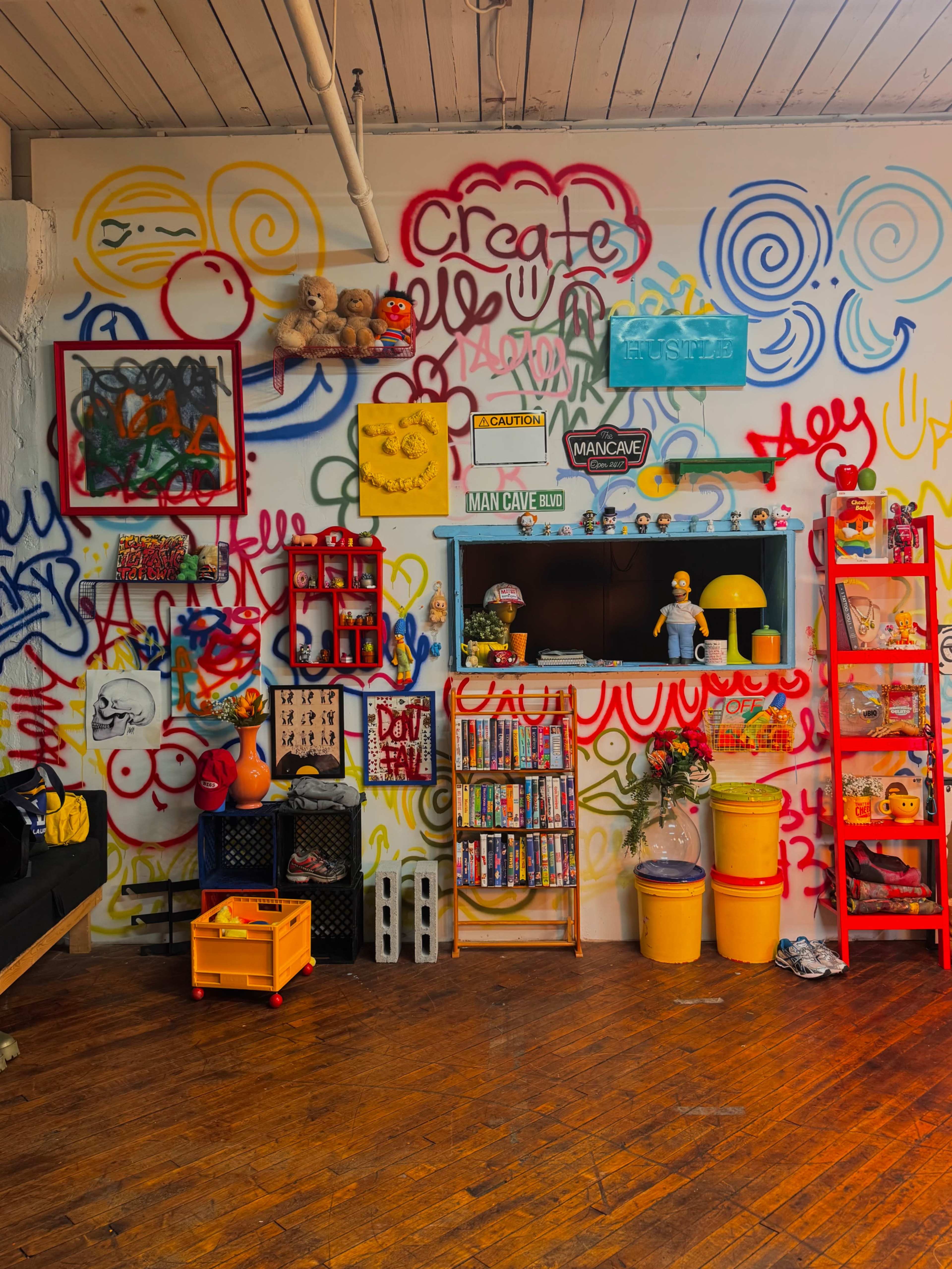 The image shows a vibrant, graffiti-covered wall with various colorful decorations, shelves, and storage items arranged in an eclectic manner.