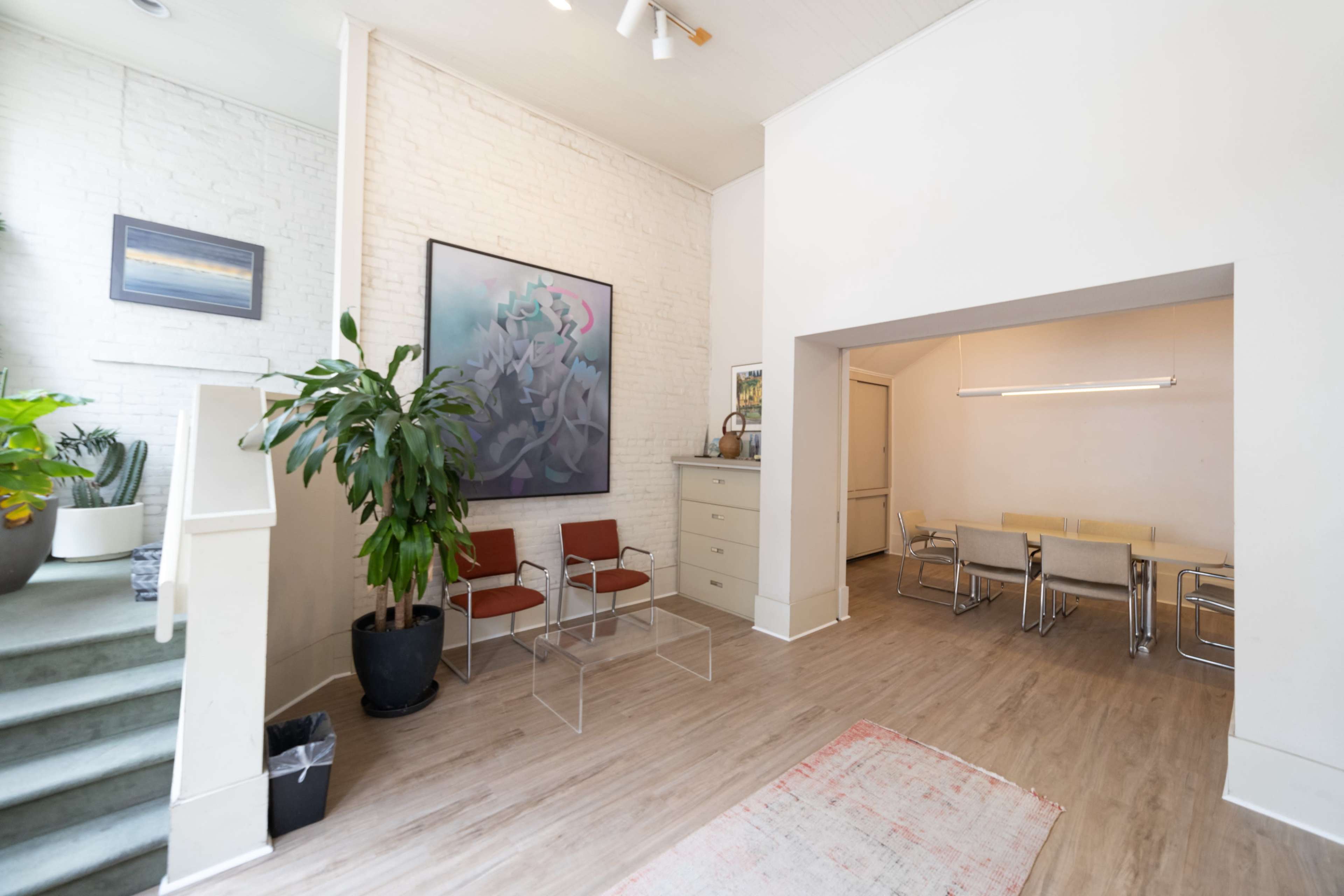A modern, airy interior space with a staircase leading to a brightly lit area featuring minimalist furniture and a large piece of art on the wall.
