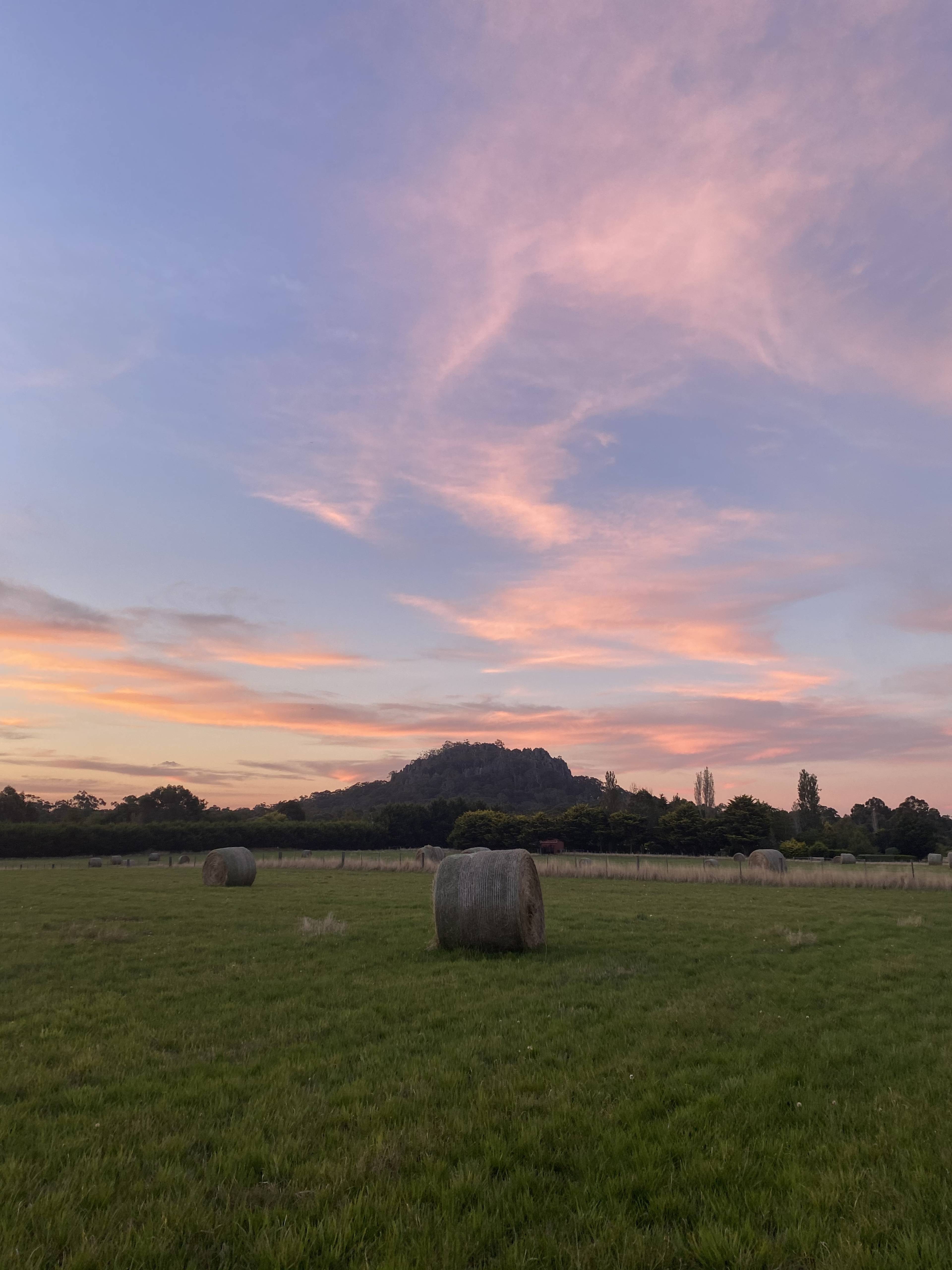 The image shows a grassy field with a hay bale in the foreground and a mountain under a colorful sky at sunset in the background.