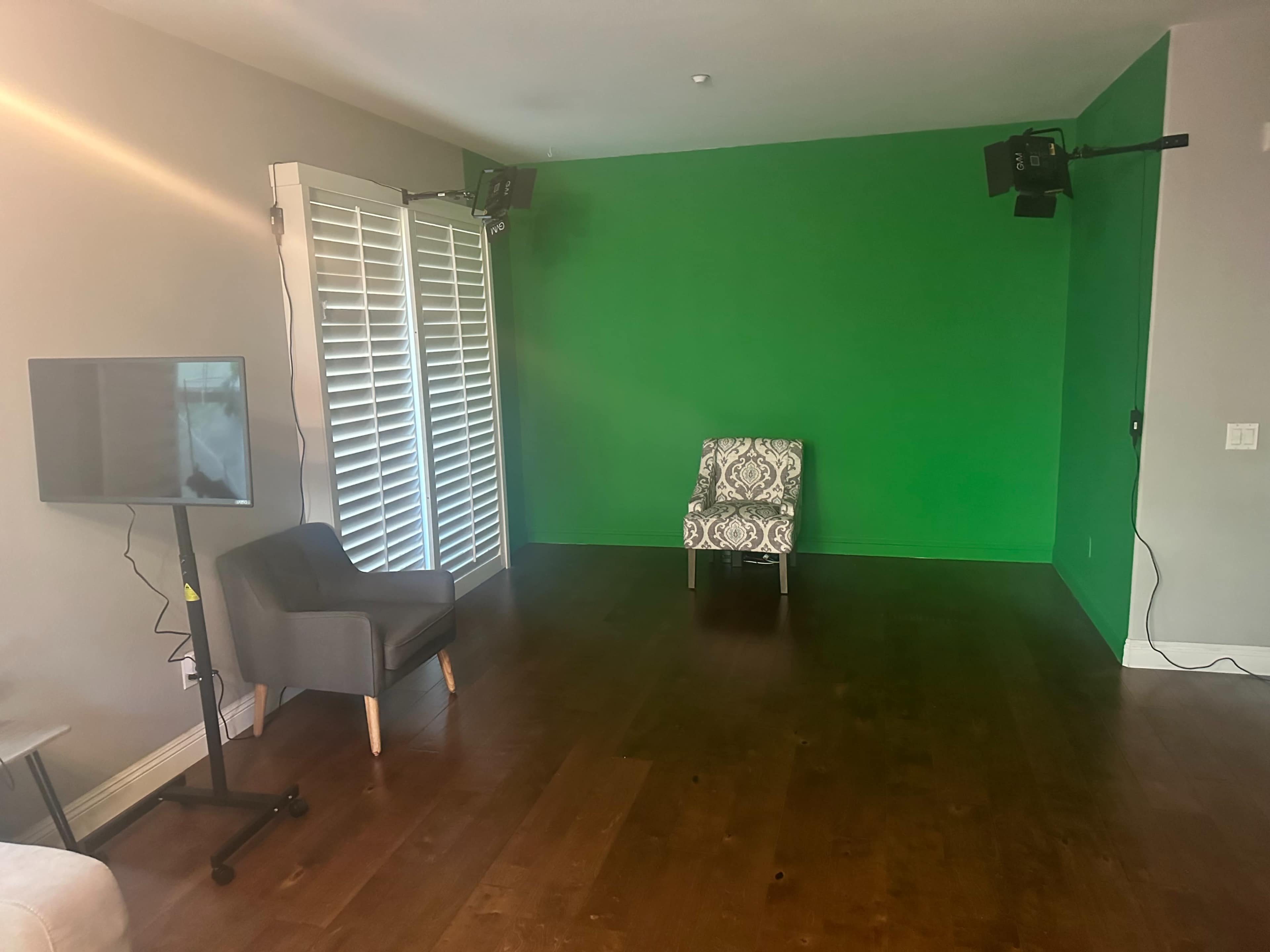 A vacant room with a green screen backdrop, a patterned chair, a television on a stand, and wooden flooring.