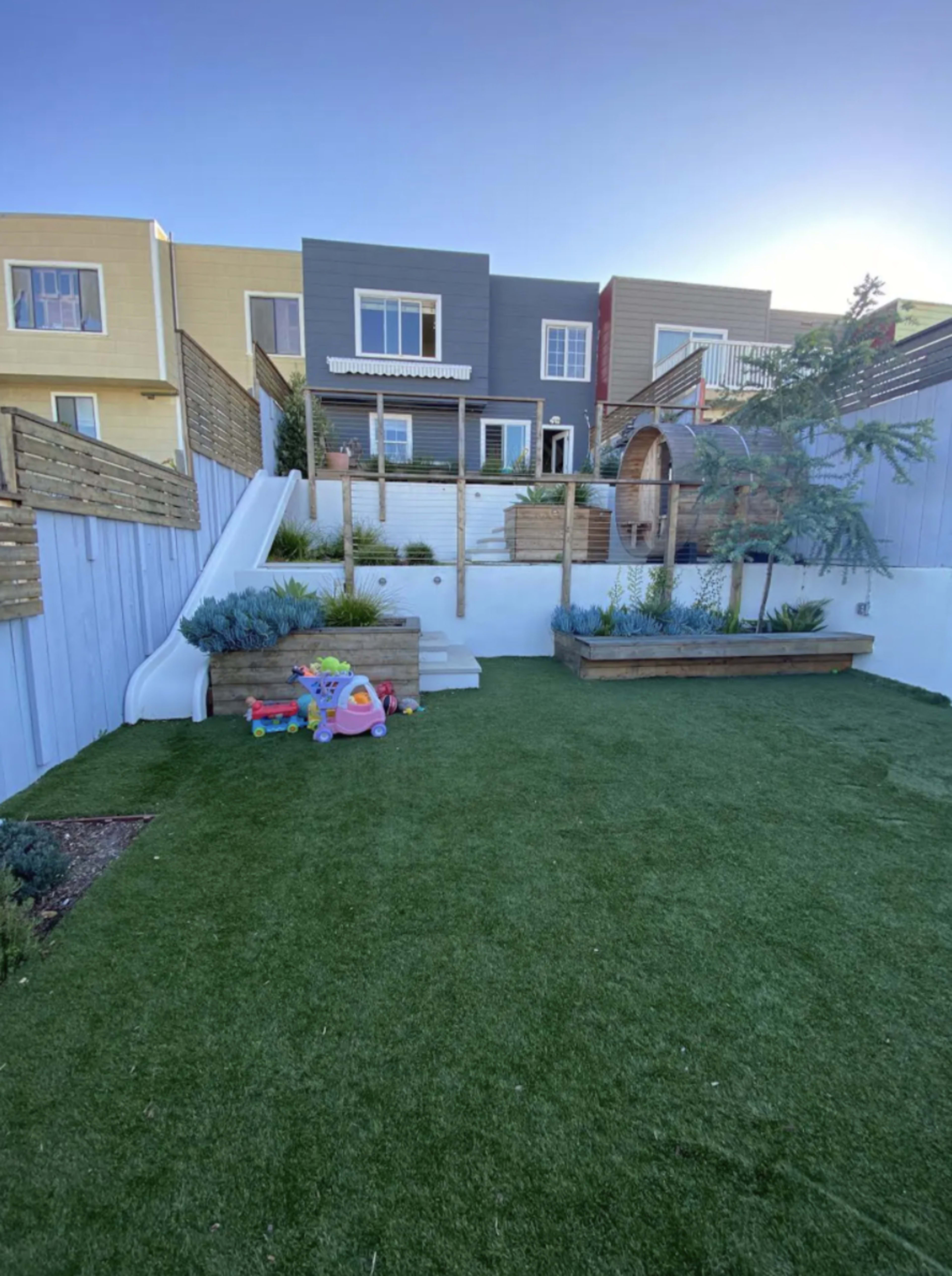 The image shows a backyard with artificial grass, a wooden deck, planted greenery, and a child's toy set in the corner.