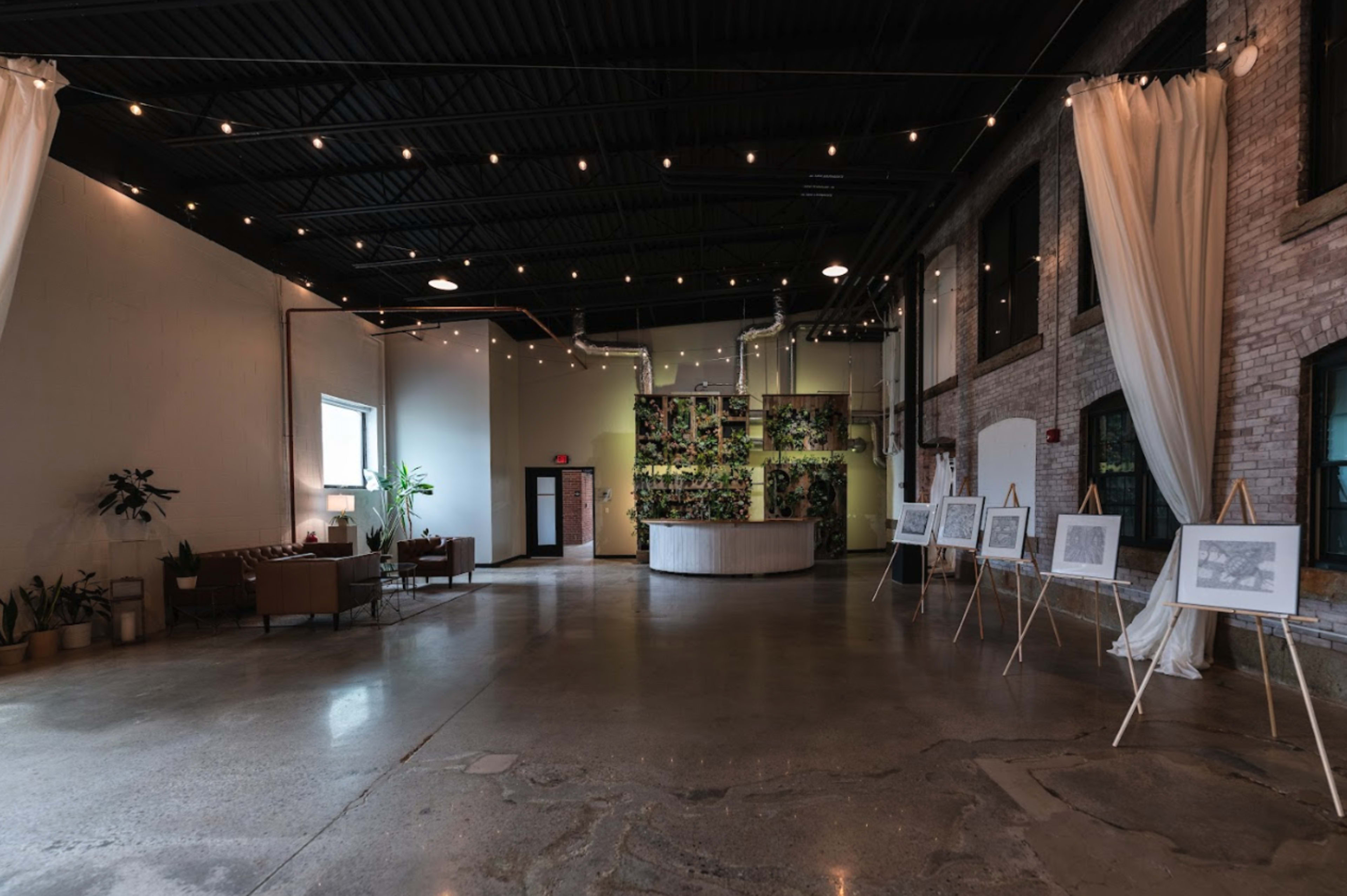 The image shows a spacious, industrial-style interior with a seating area, potted plants, and framed artwork displayed on easels along the walls.