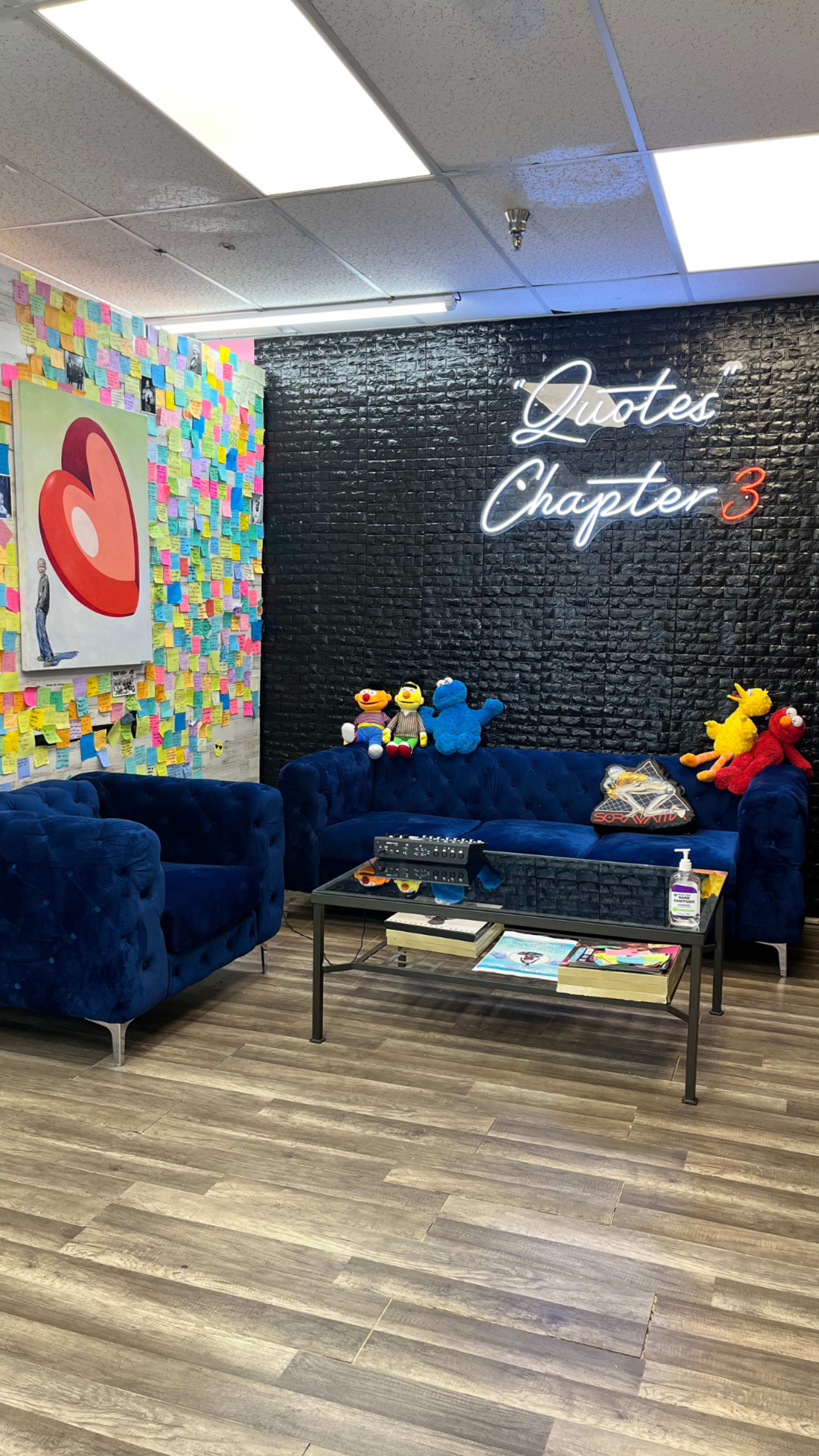 A cozy lounge area with blue sofas, a glass coffee table, colorful sticky notes on the wall, and a neon sign that reads "Quotes Chapter 3."