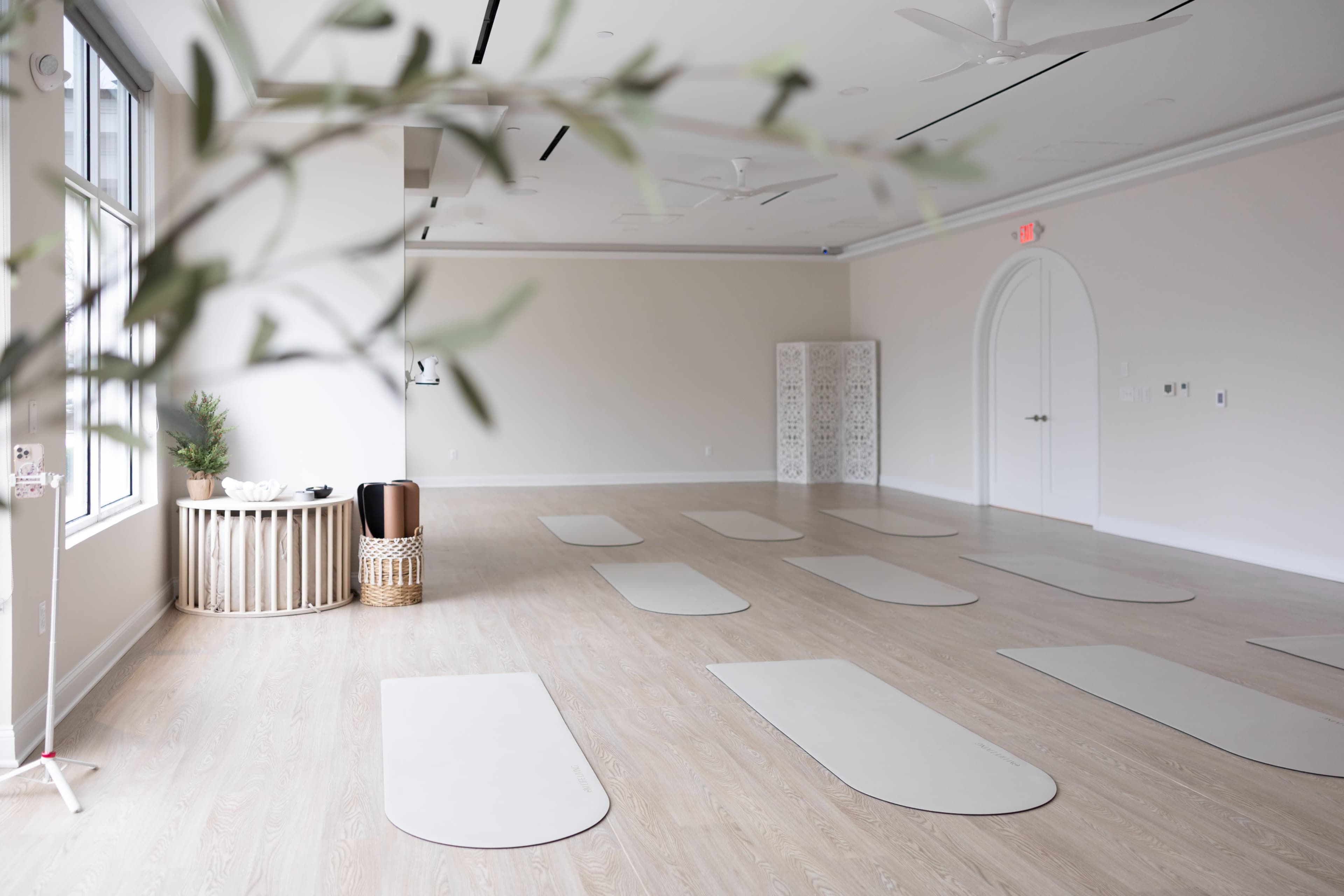 The image shows a spacious, empty yoga studio with light wooden flooring and several yoga mats arranged neatly on the floor.