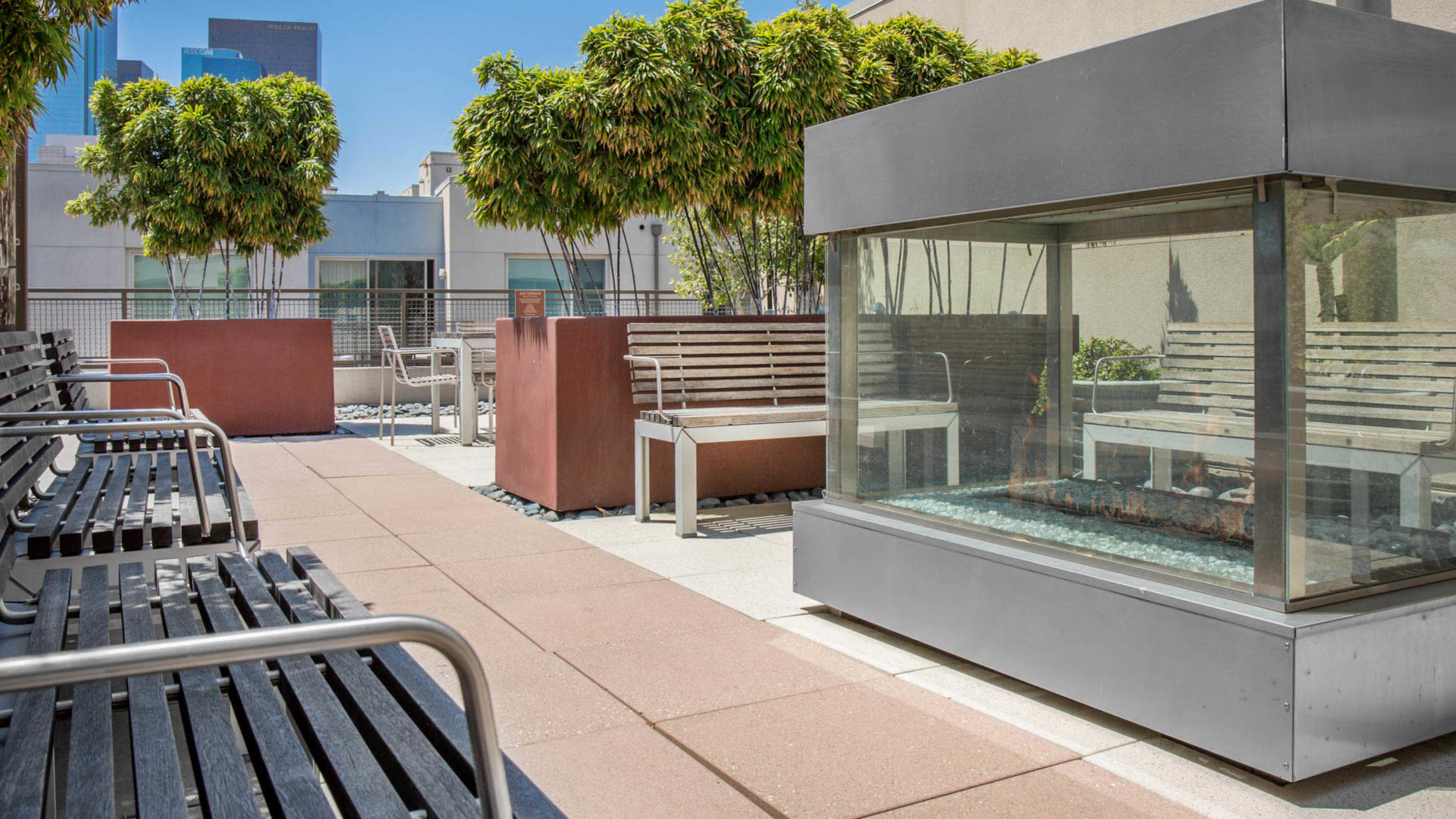 The scene features a modern urban outdoor lounge area with benches, planters, and a glass-enclosed fire pit surrounded by landscaping.
