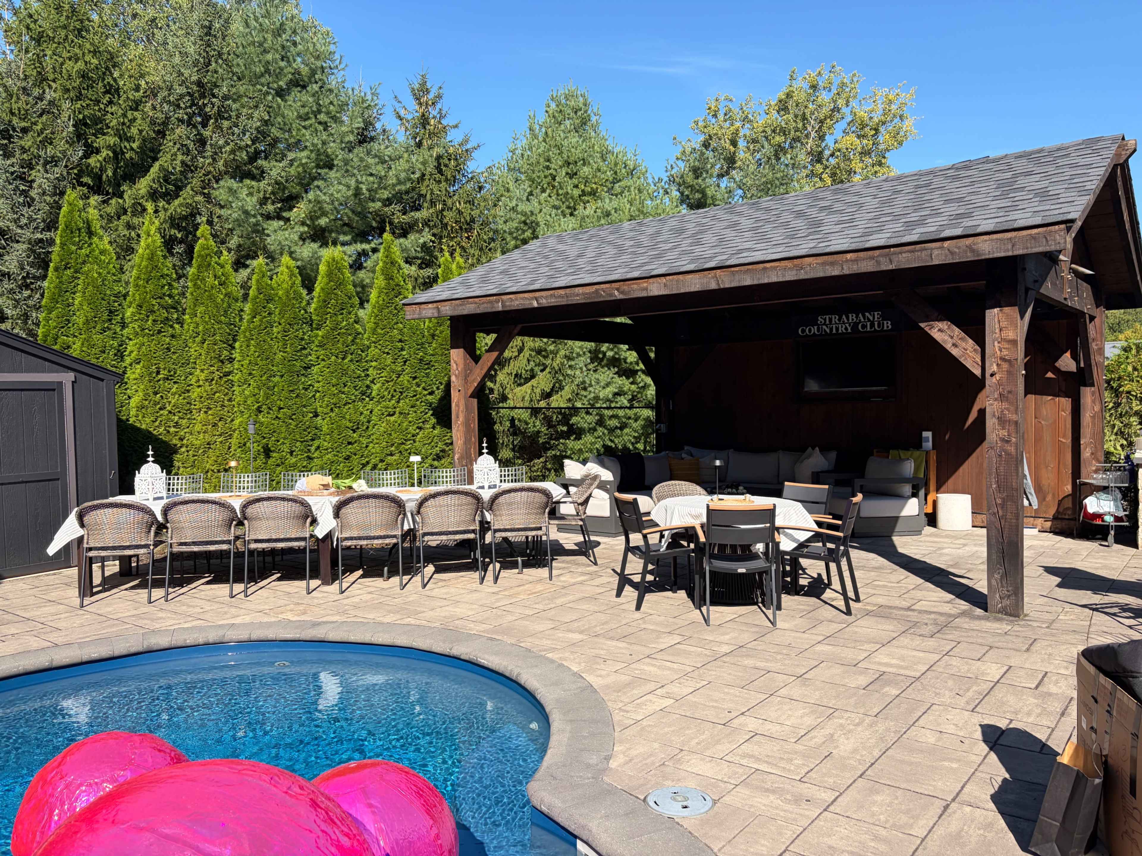 The image shows an outdoor pool area with a patio featuring ample seating and a covered lounge space surrounded by greenery.
