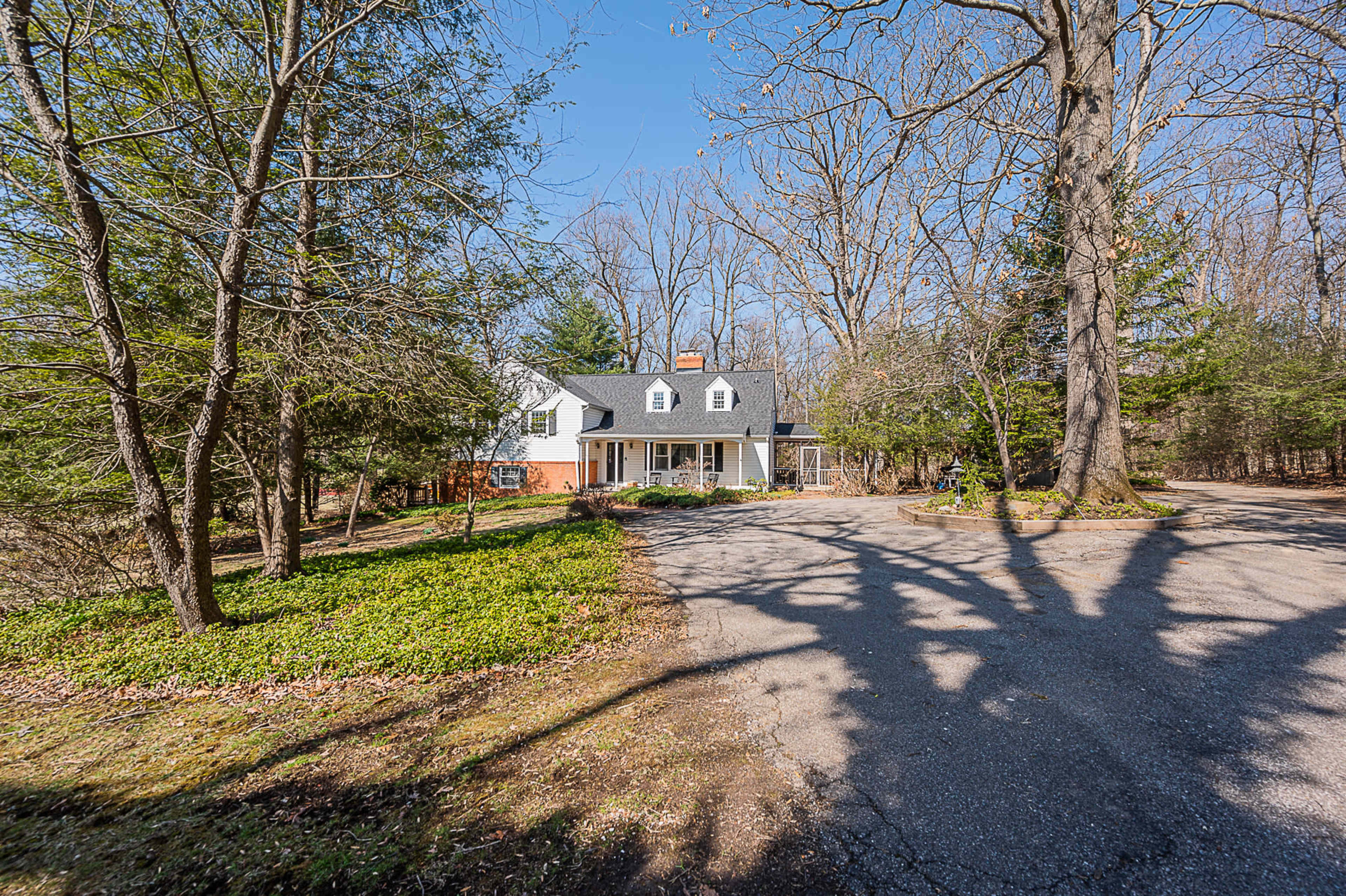A two-story house with a gabled roof is situated on a wooded property with a circular driveway surrounded by leafless trees.