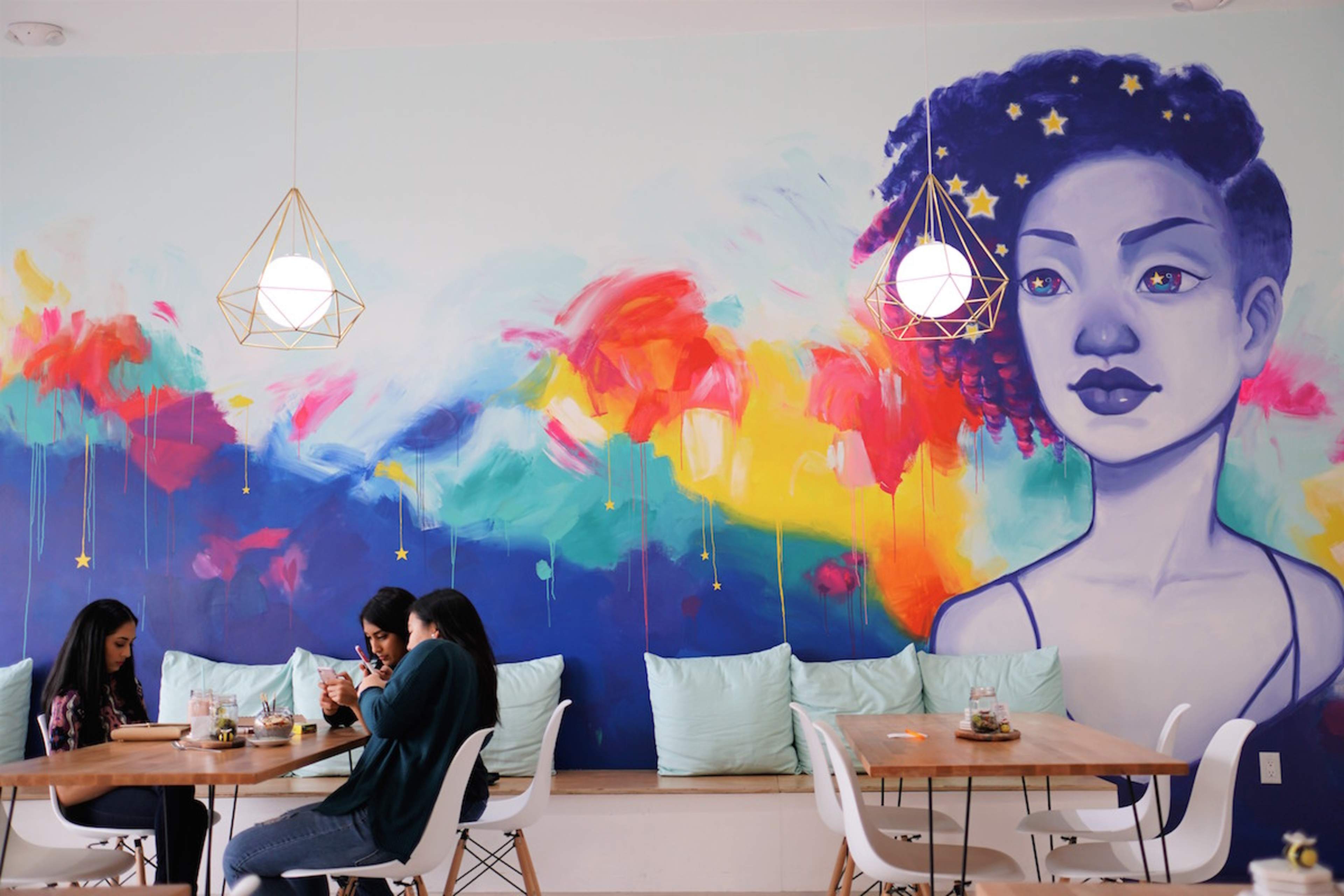 A colorful mural of a woman adorns the wall above a group of three women seated at a wooden table in a cafe.
