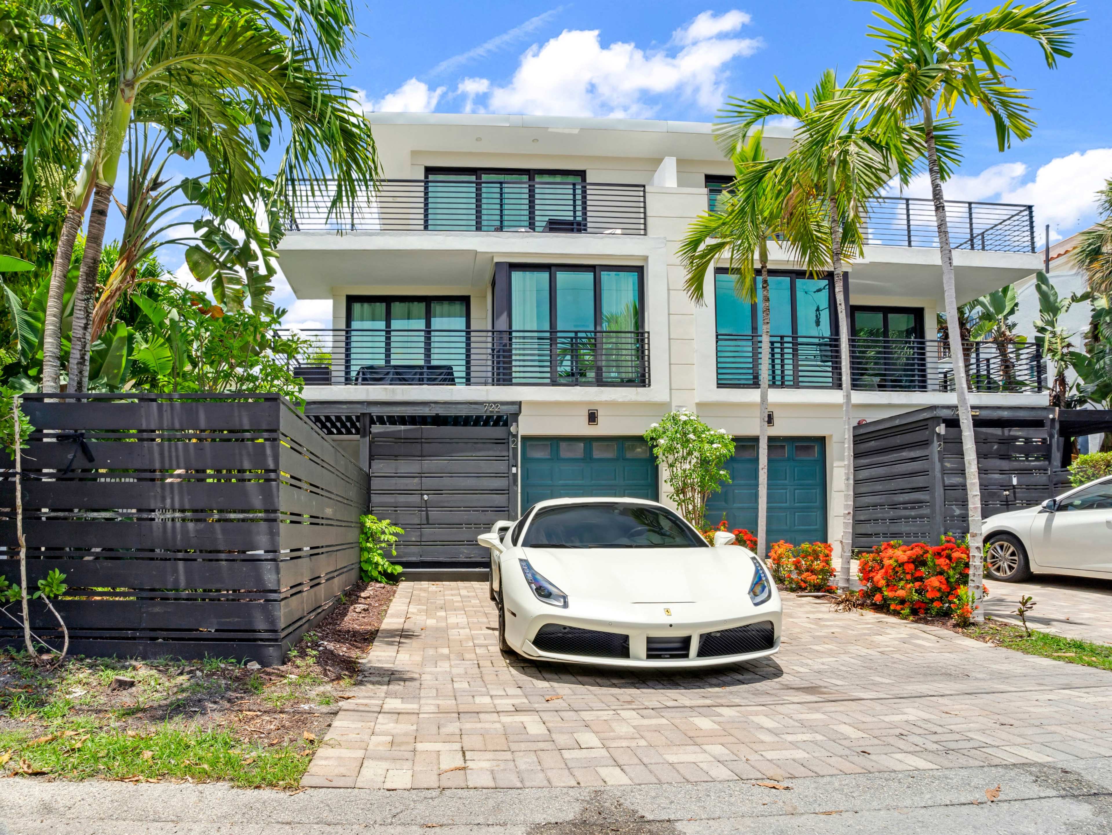 A modern three-story house with large windows is situated in front of a paved driveway, where a white sports car is parked alongside tropical plants and flowers.