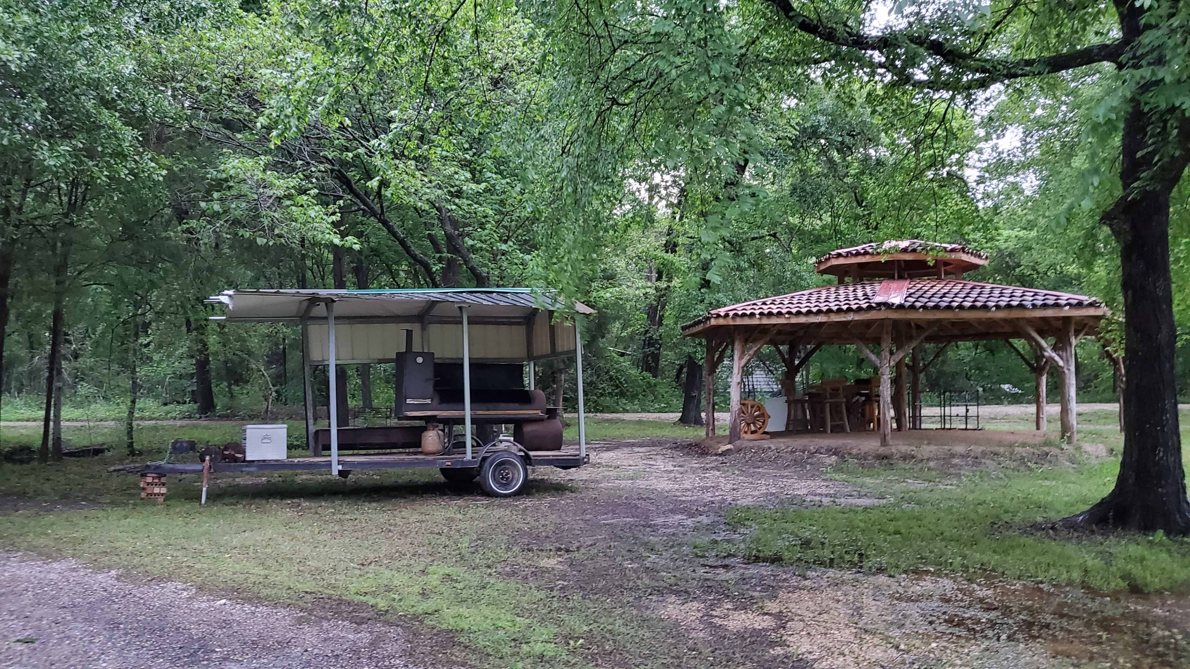 A trailer with a covered cooking area is parked next to a wooden gazebo surrounded by trees in a grassy area.
