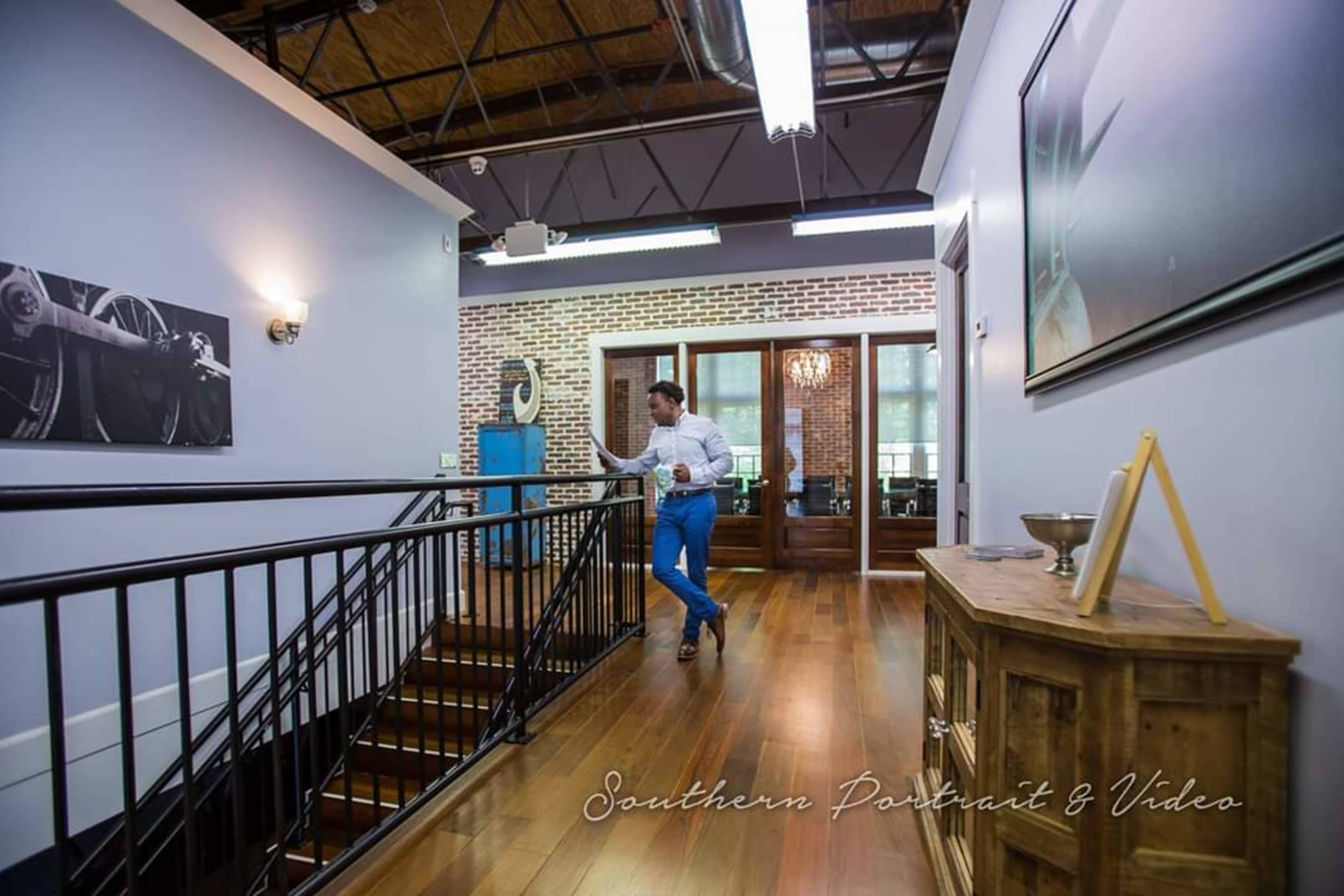 A man in a white shirt and blue trousers walks along a wooden hallway with a railing, passing by brick walls and doorways.