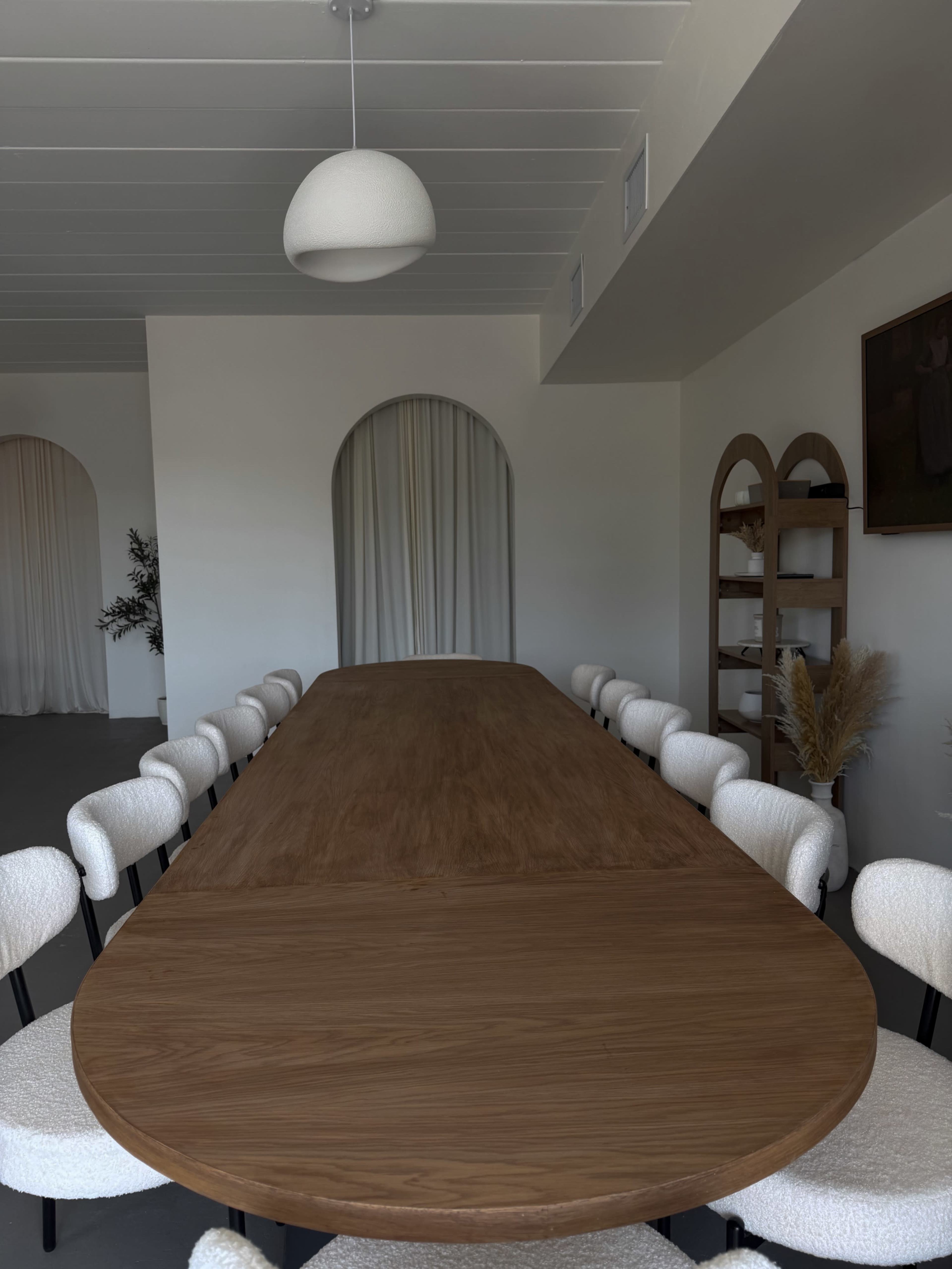 A large, oval wooden dining table is surrounded by multiple white chairs in a well-lit room featuring a simple pendant light and an arched doorway.