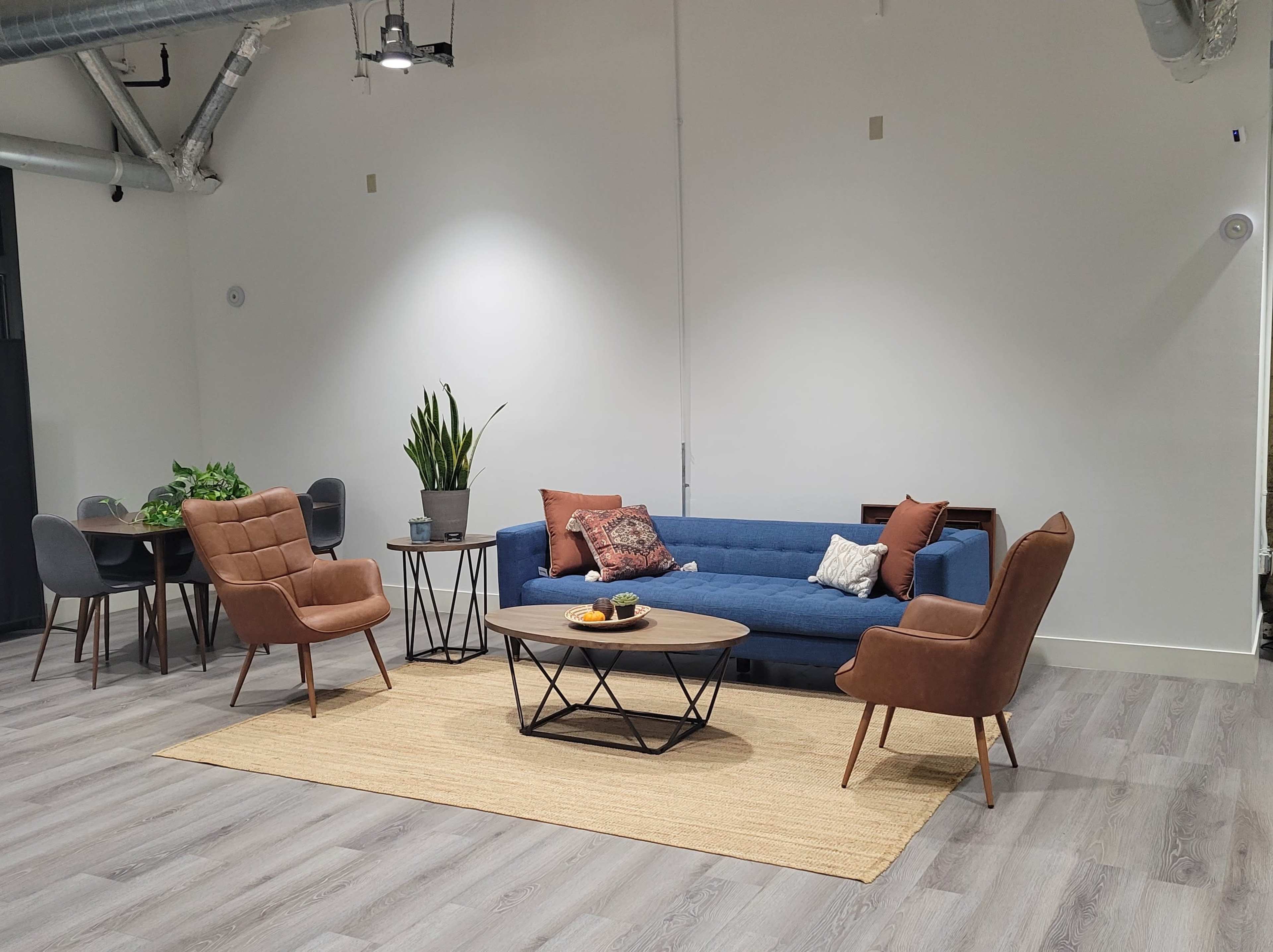 A modern living room features a blue couch, two brown armchairs, a round coffee table with fruit, a decorative rug, and a dining area with a plant.