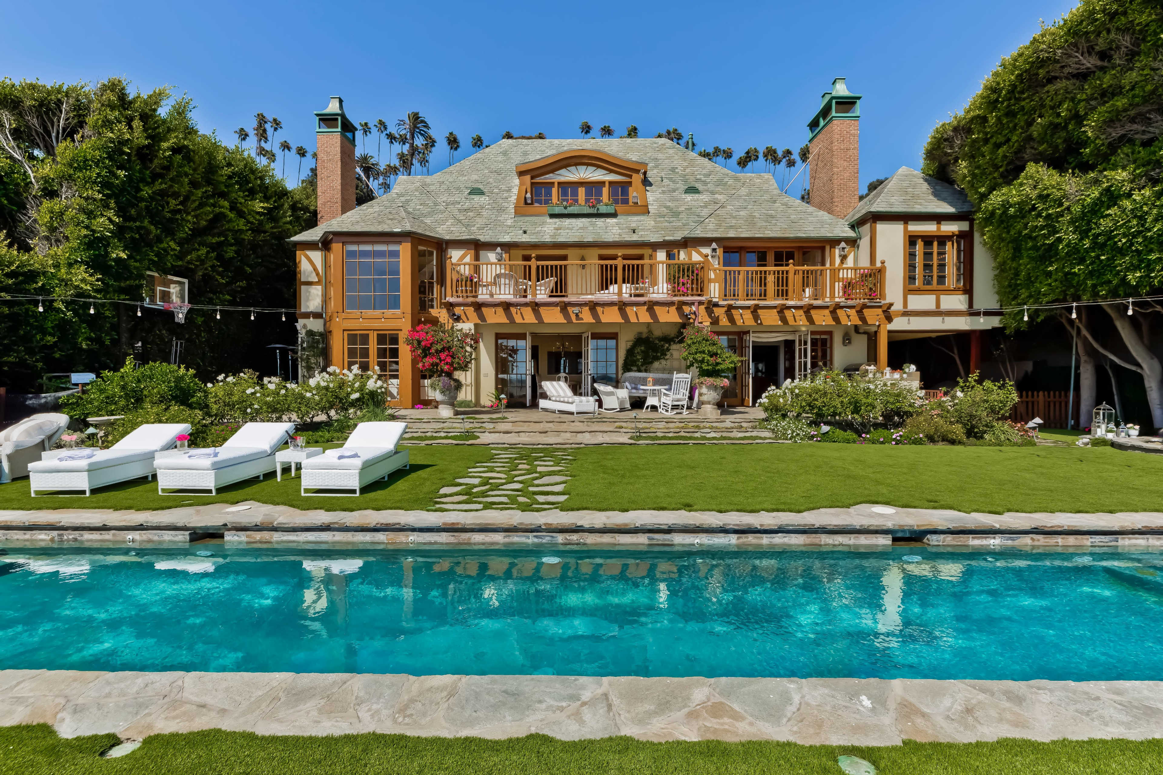 The image shows a large, two-story house with a manicured lawn and a swimming pool in the foreground, surrounded by greenery and outdoor seating areas.