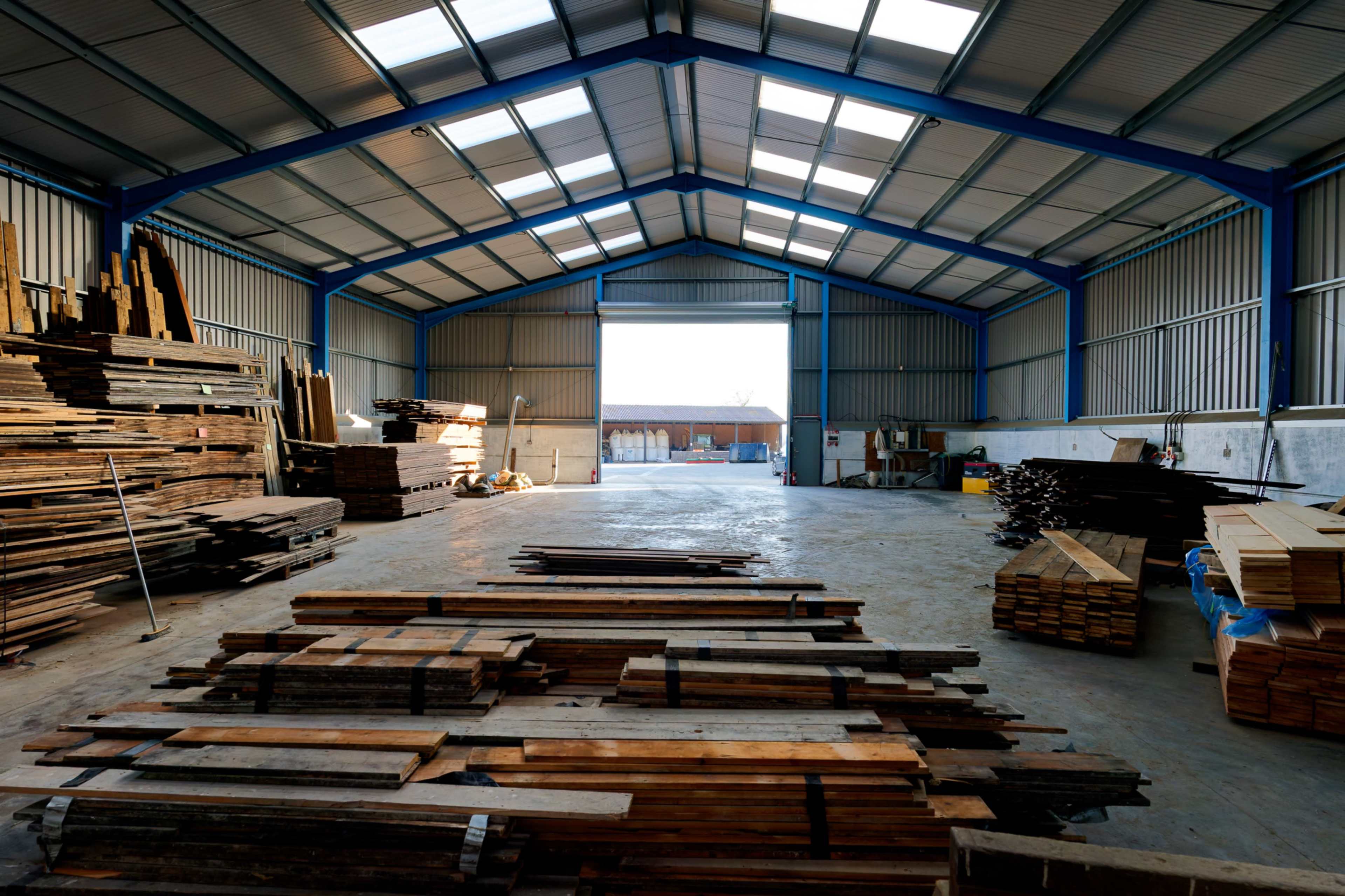 The image shows an interior view of a spacious warehouse with stacks of wooden planks and beams arranged along the walls.