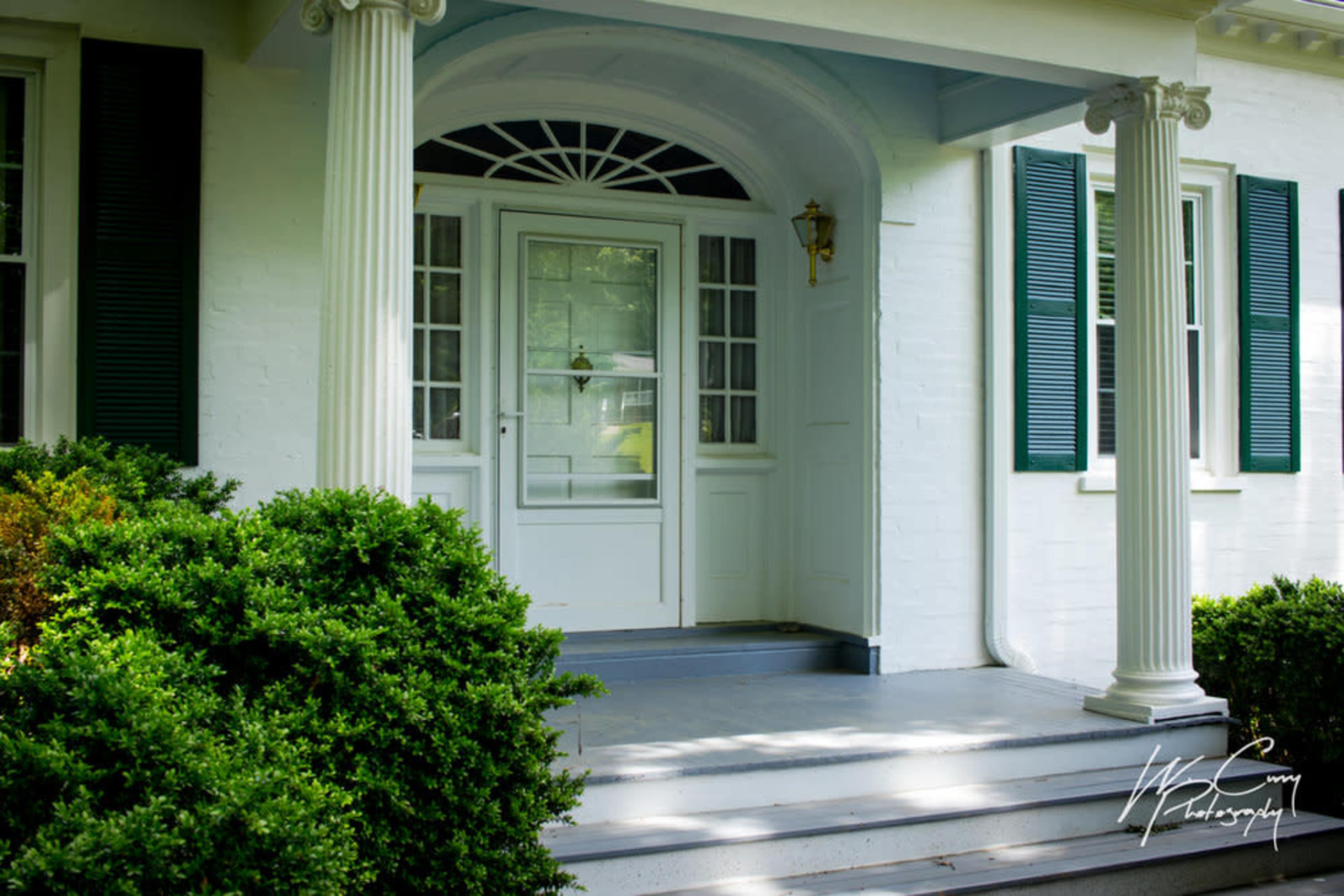 A front porch with white columns, a front door, and green shrubs.
