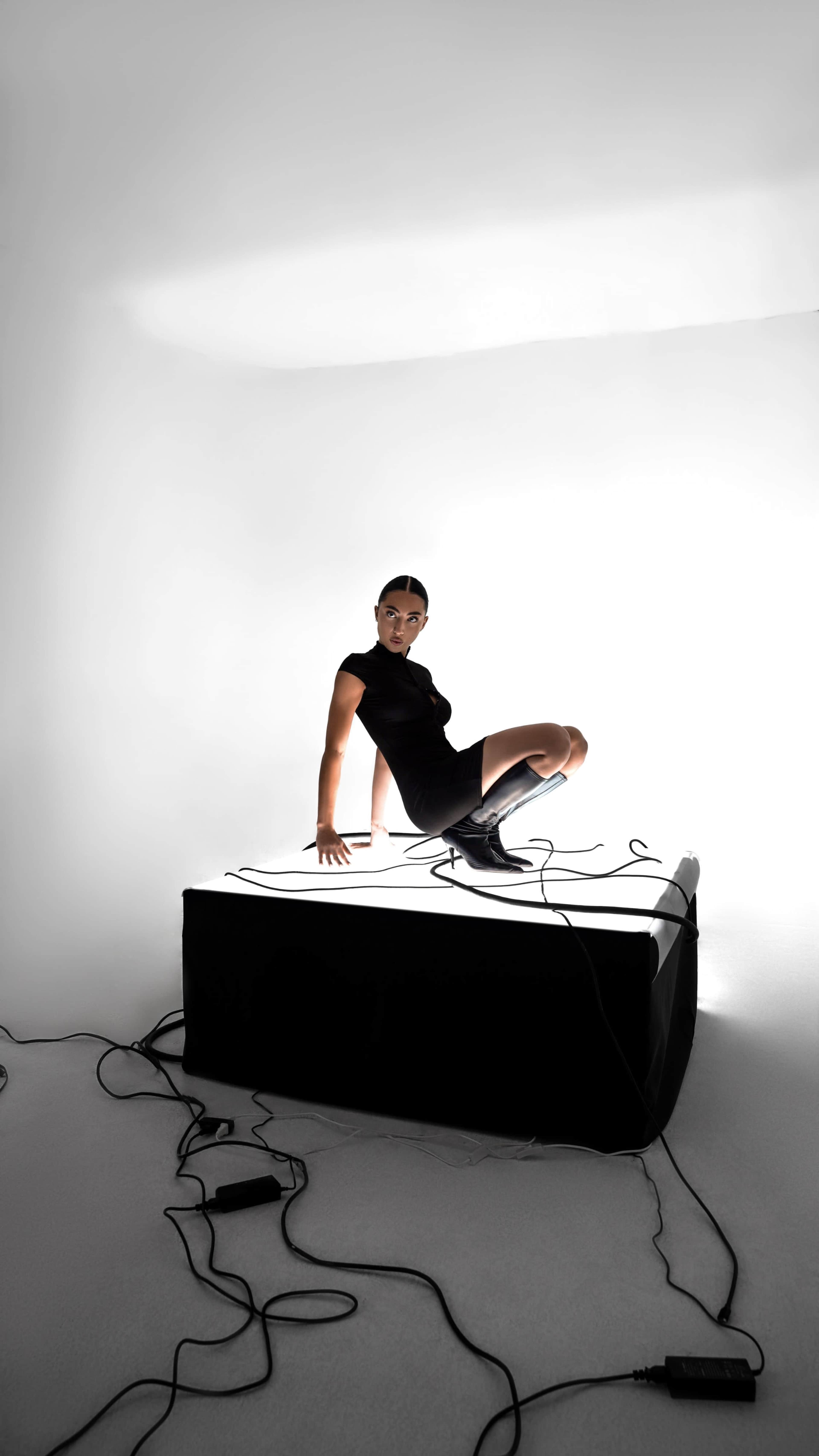 A person in a black outfit is seated on a lighted platform with various cables around it.
