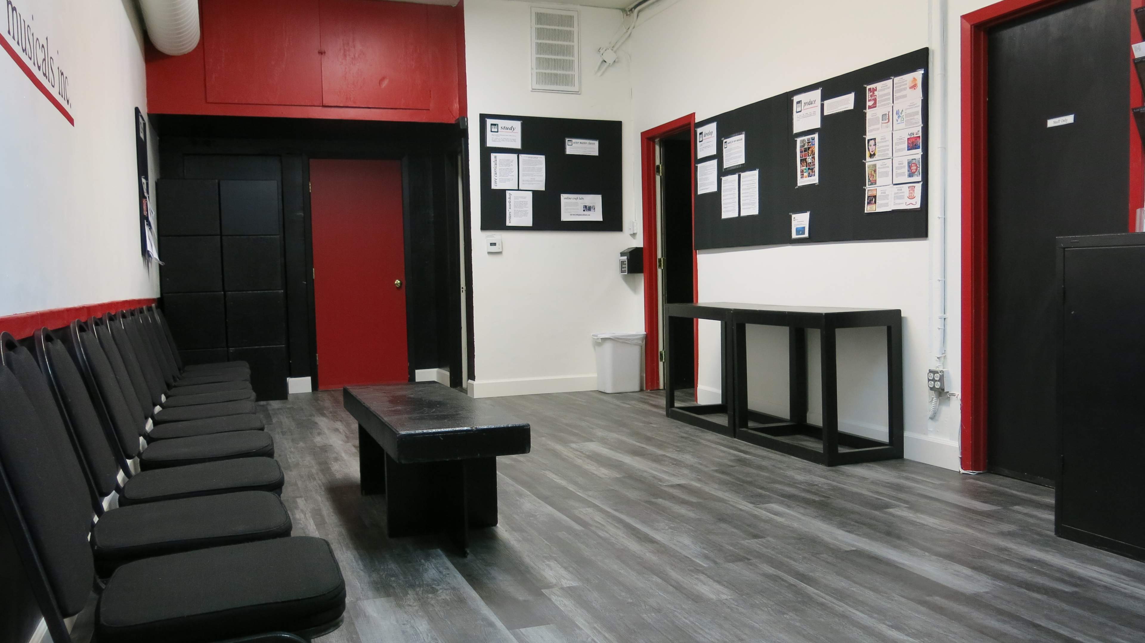 The image shows a waiting area with a row of black chairs, a long black table, and bulletin boards on the walls.