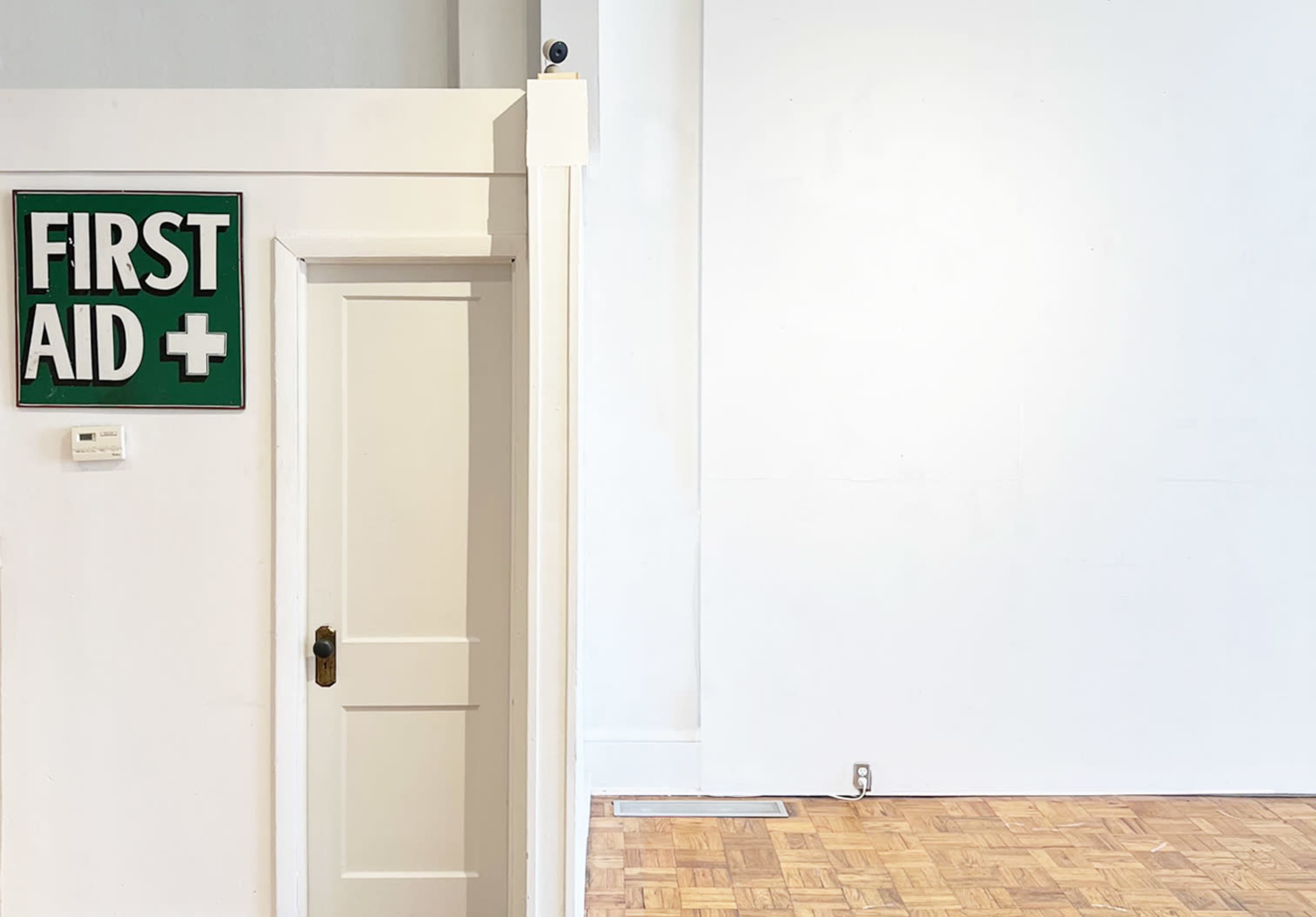 A white-walled room features a clear doorway labeled "FIRST AID" next to an empty space with parquet flooring.