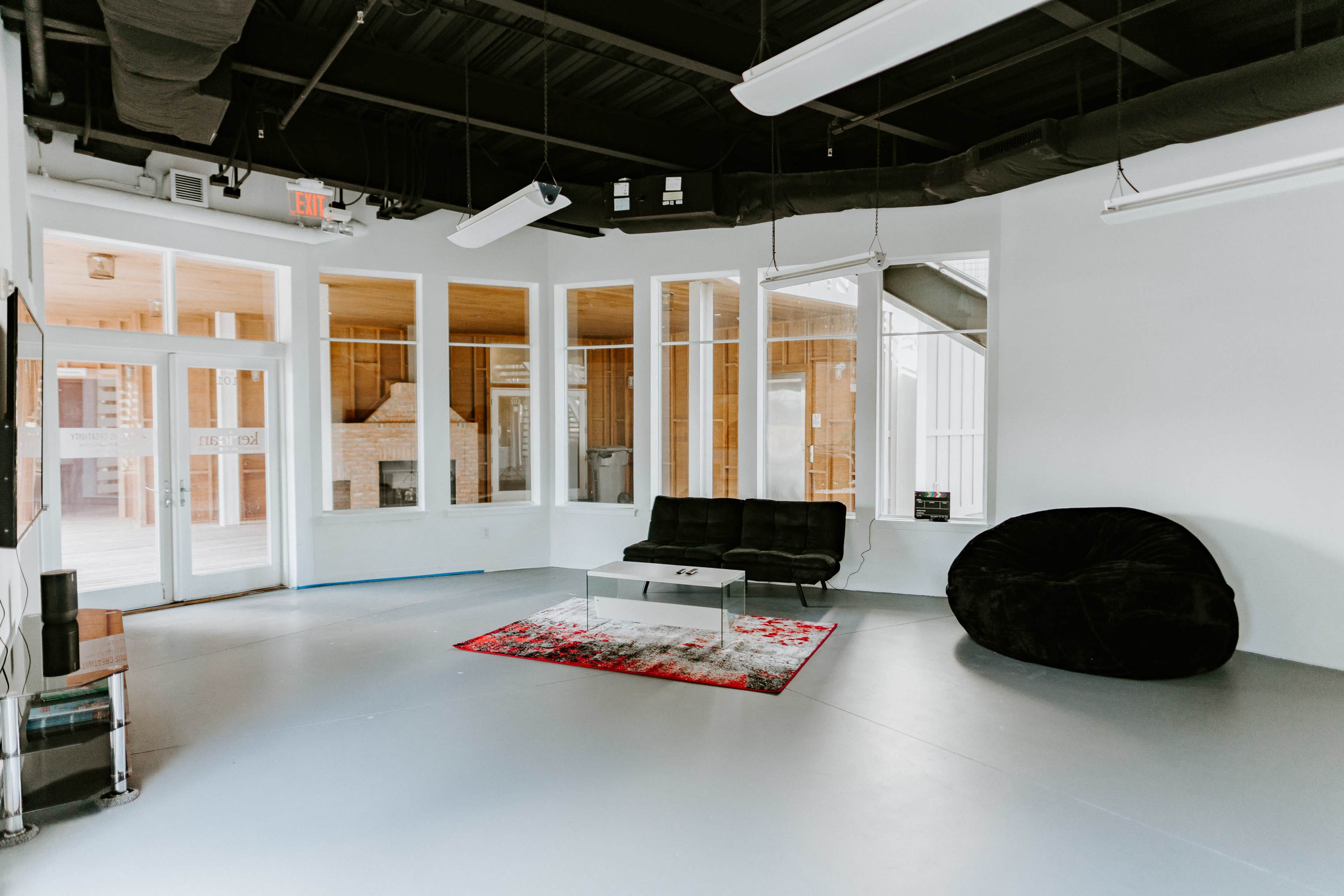 A modern, open space with a black sofa, a large bean bag chair, and a glass coffee table on a colorful rug, surrounded by large windows.