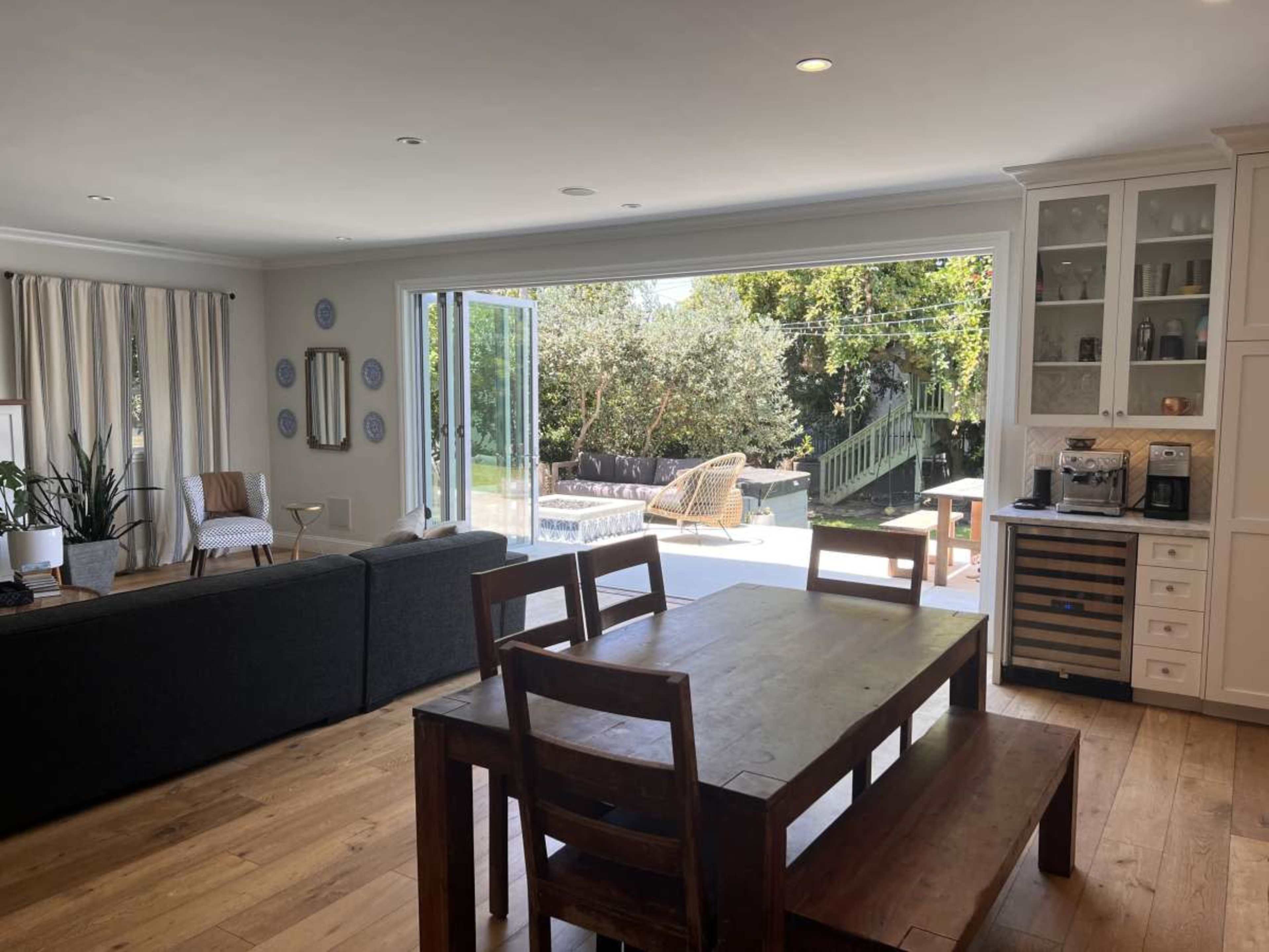 The image shows an open living and dining area with a wooden table, a gray sofa, and large sliding glass doors that lead to a backyard patio.
