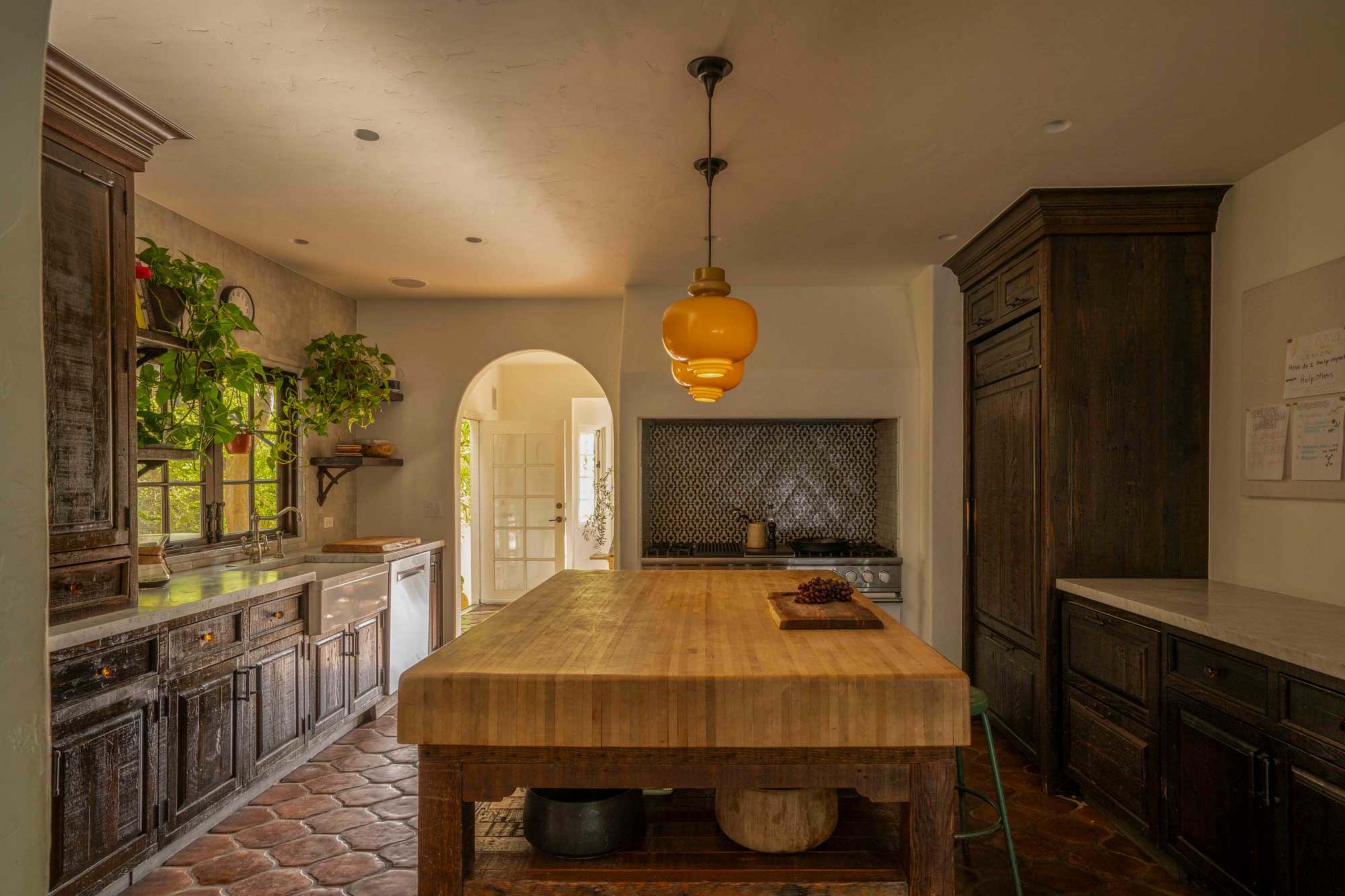 A spacious kitchen features a large wooden island, dark cabinetry, and plants on the windowsill, with an arched doorway leading to another room.