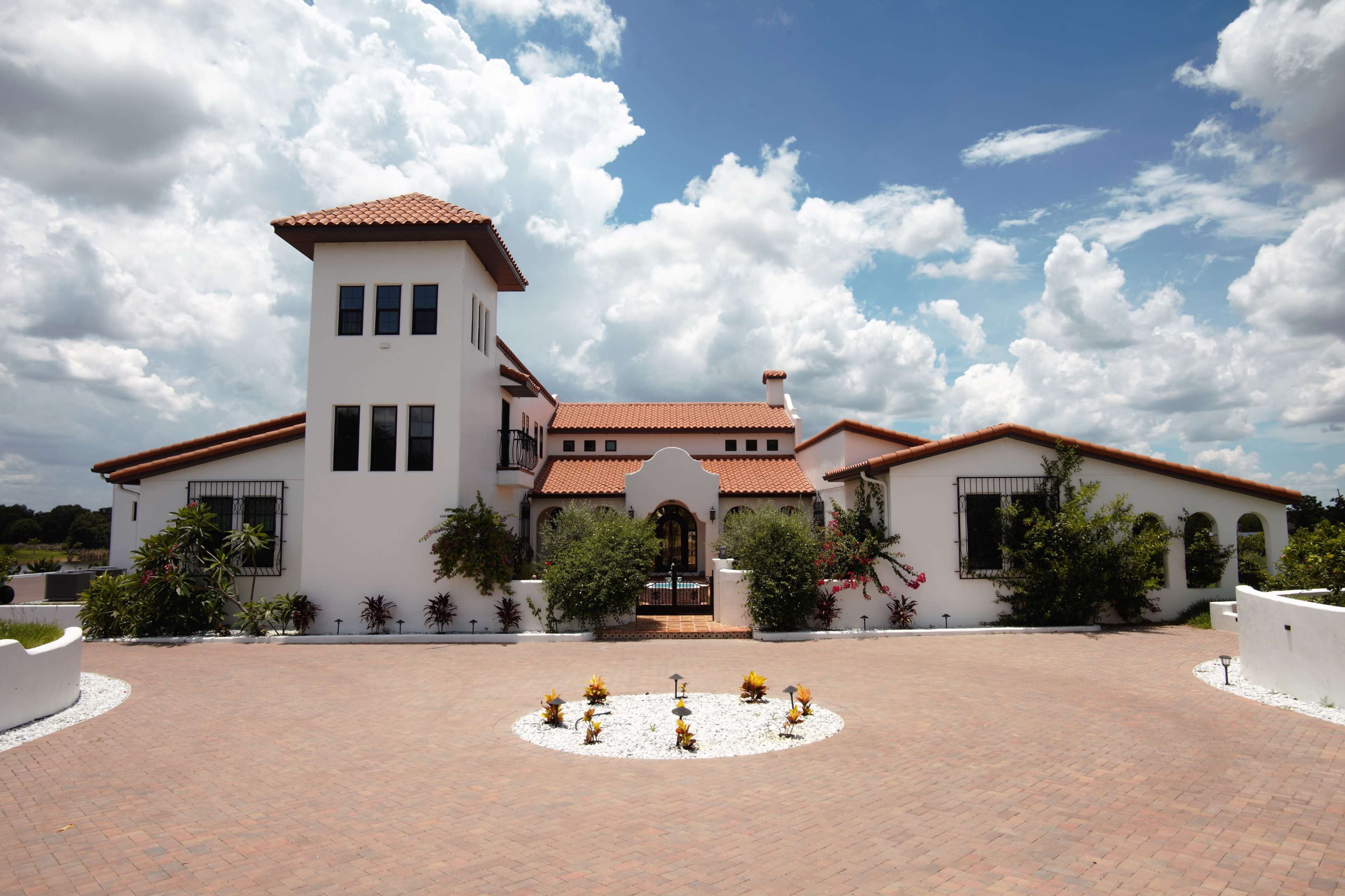 A large, white stucco house with a tiled roof features a tower and a landscaped front yard with a circular flowerbed.