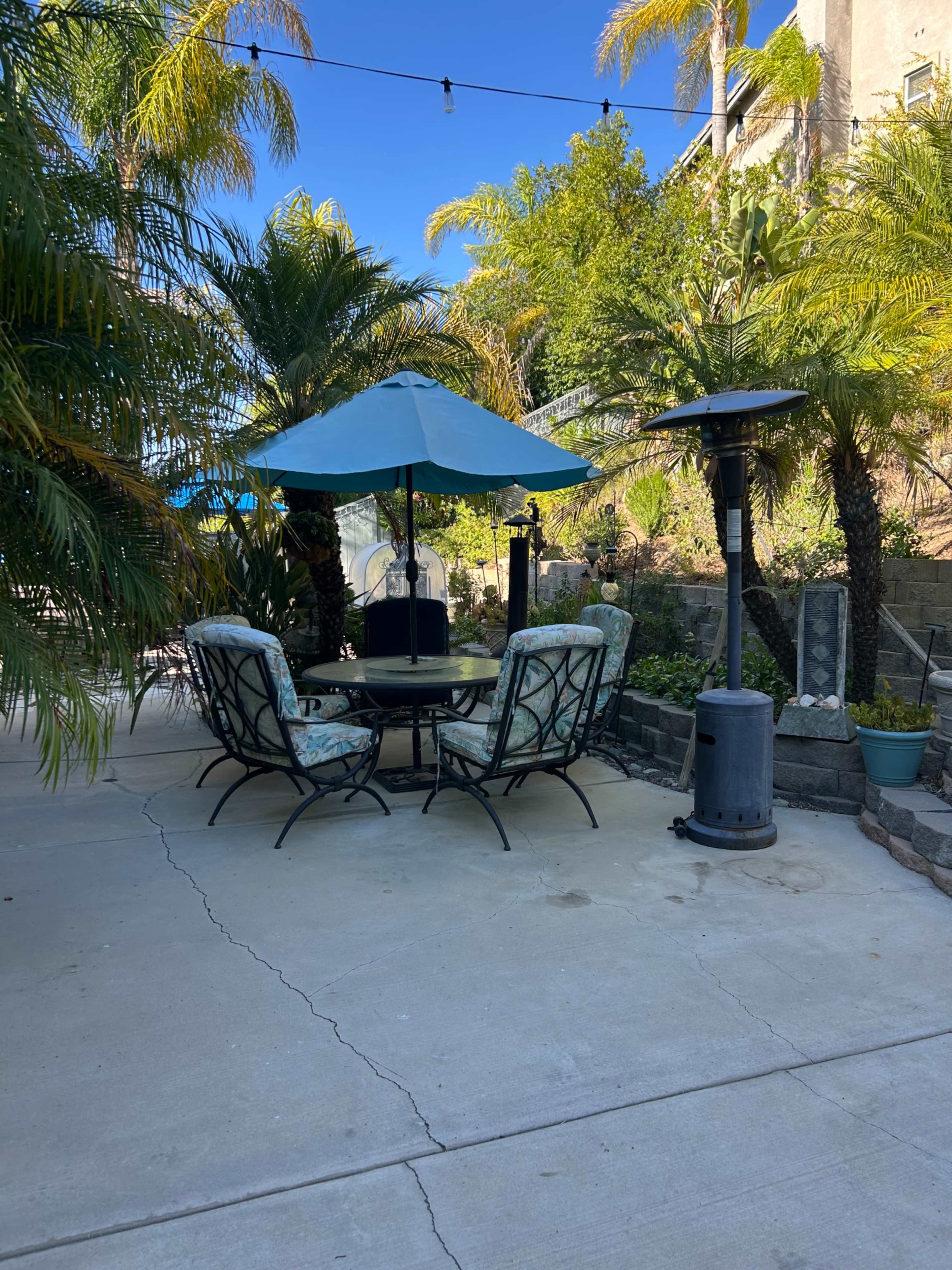 A seating area with a round table under a blue umbrella is surrounded by palm trees and outdoor heaters on a concrete patio.