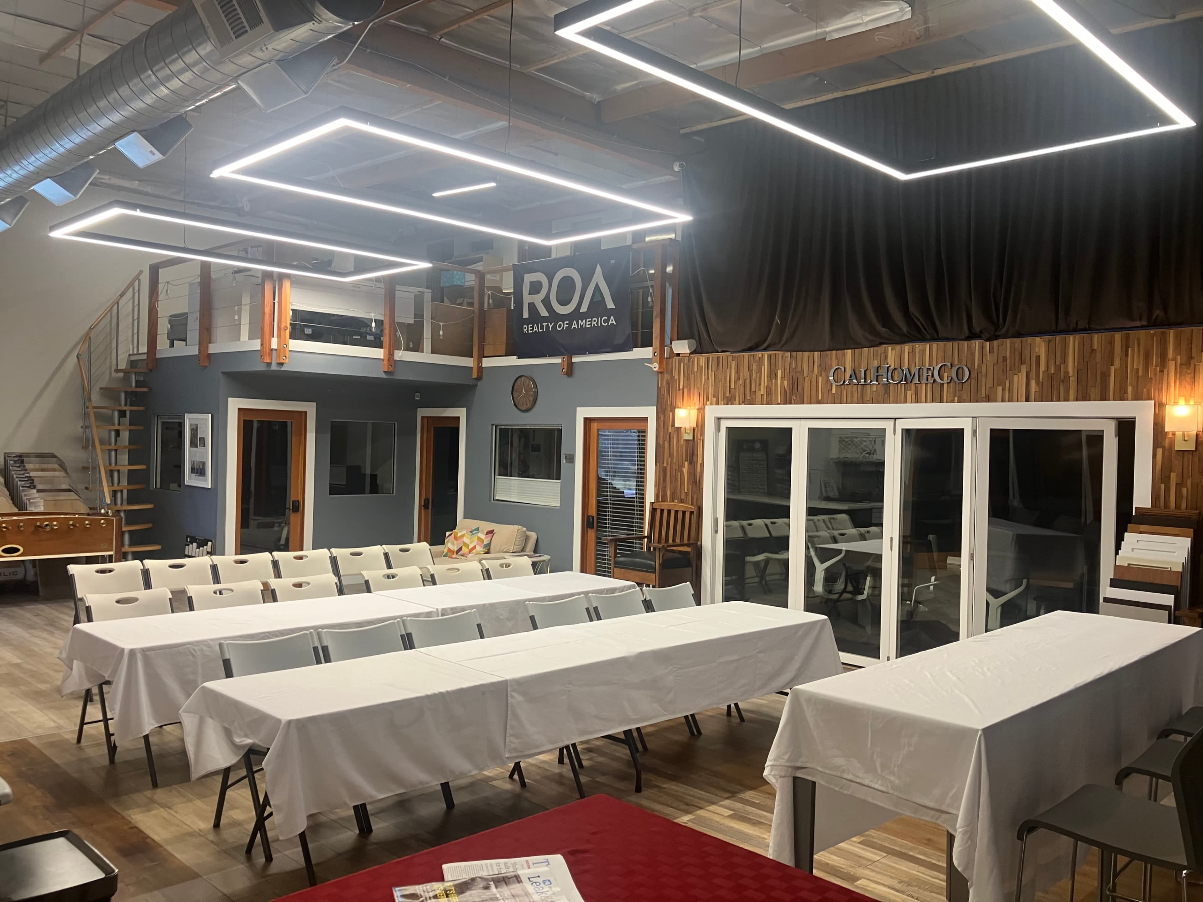 The image depicts a modern indoor meeting space with rows of white tables and chairs set up, surrounded by wooden accents and large windows.