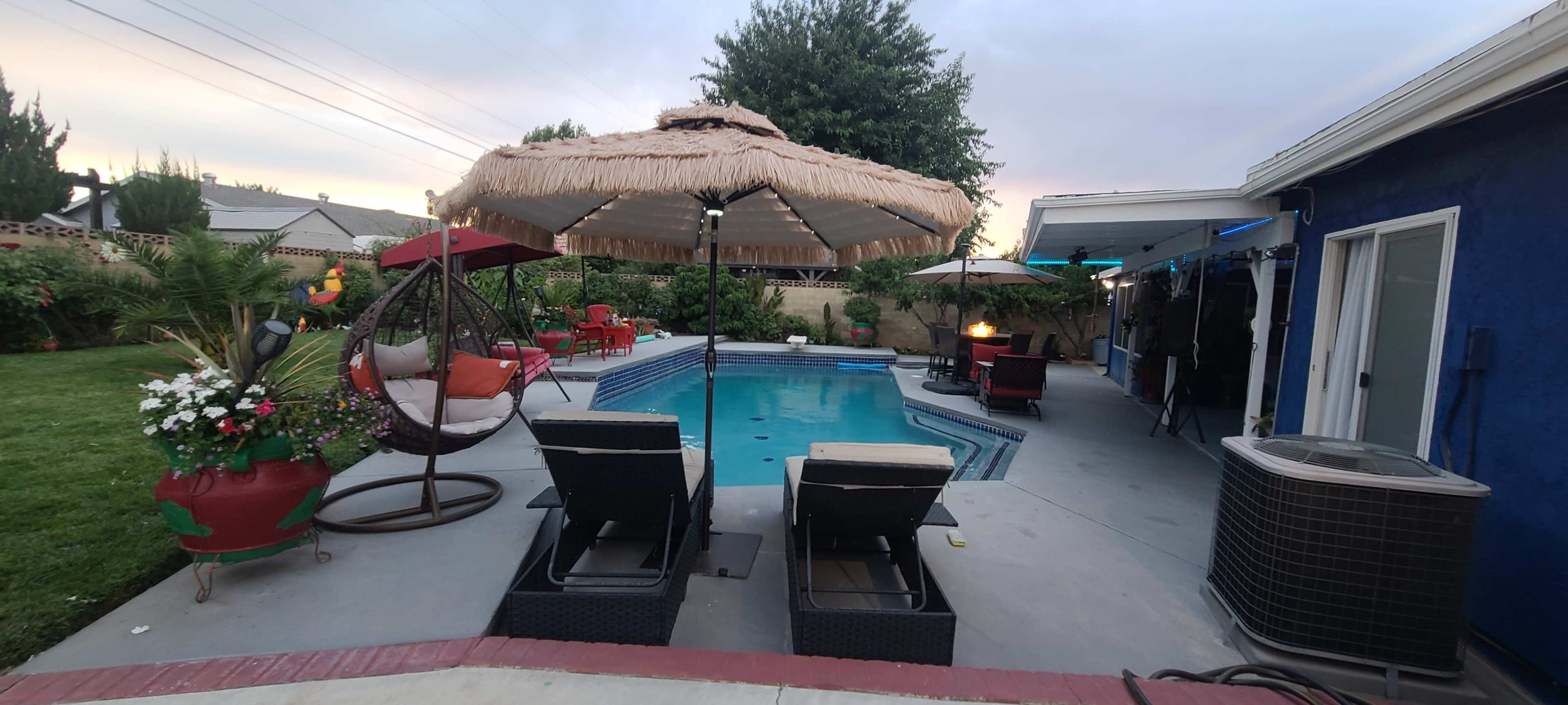 A backyard pool area featuring lounge chairs, a thatched umbrella, and a mix of patio furniture surrounded by trees and garden plants.