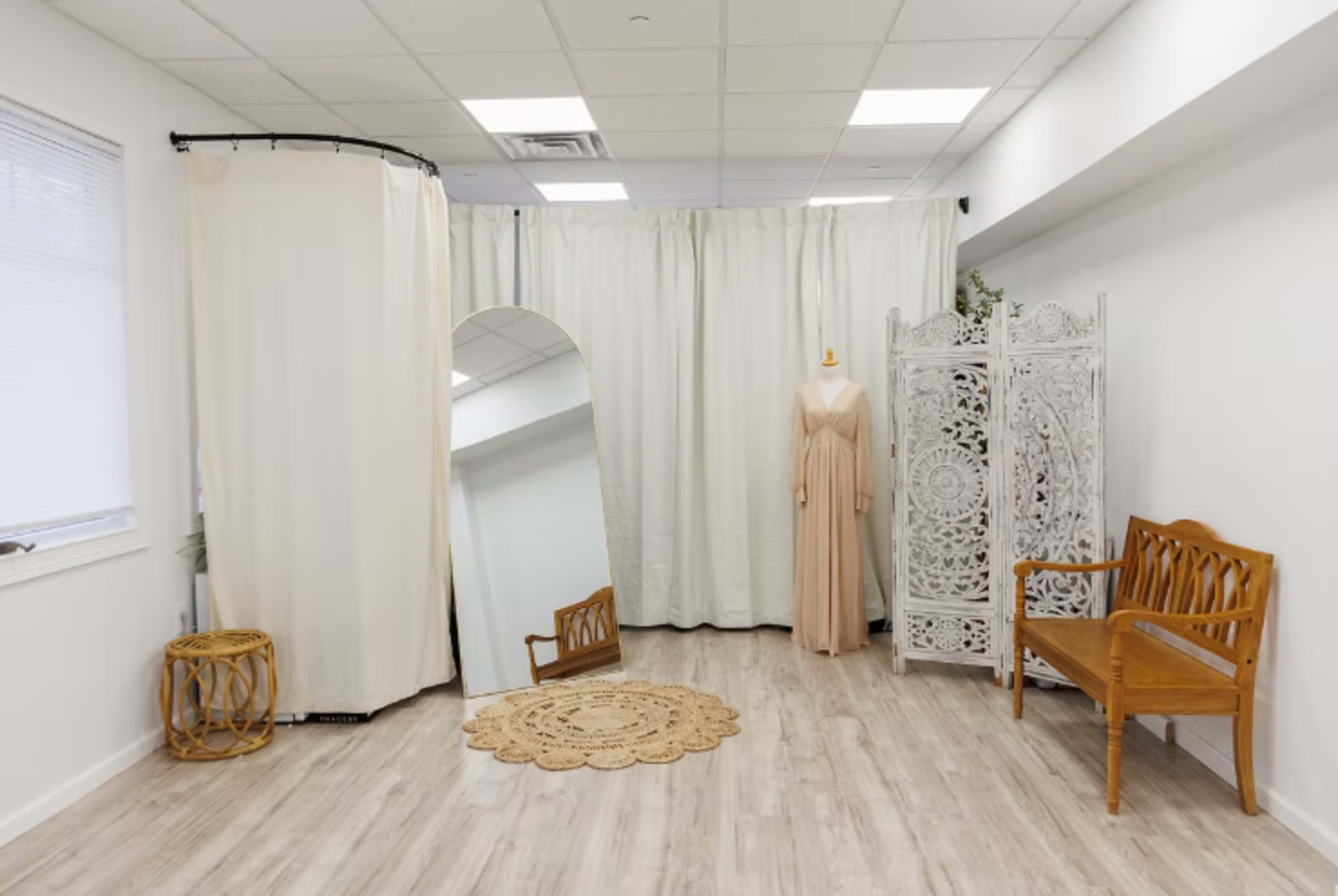 The image shows a spacious interior with a beige color scheme featuring a curved curtain divider, a full-length mirror, a mannequin dressed in a long gown, a decorative screen, and wooden furniture.