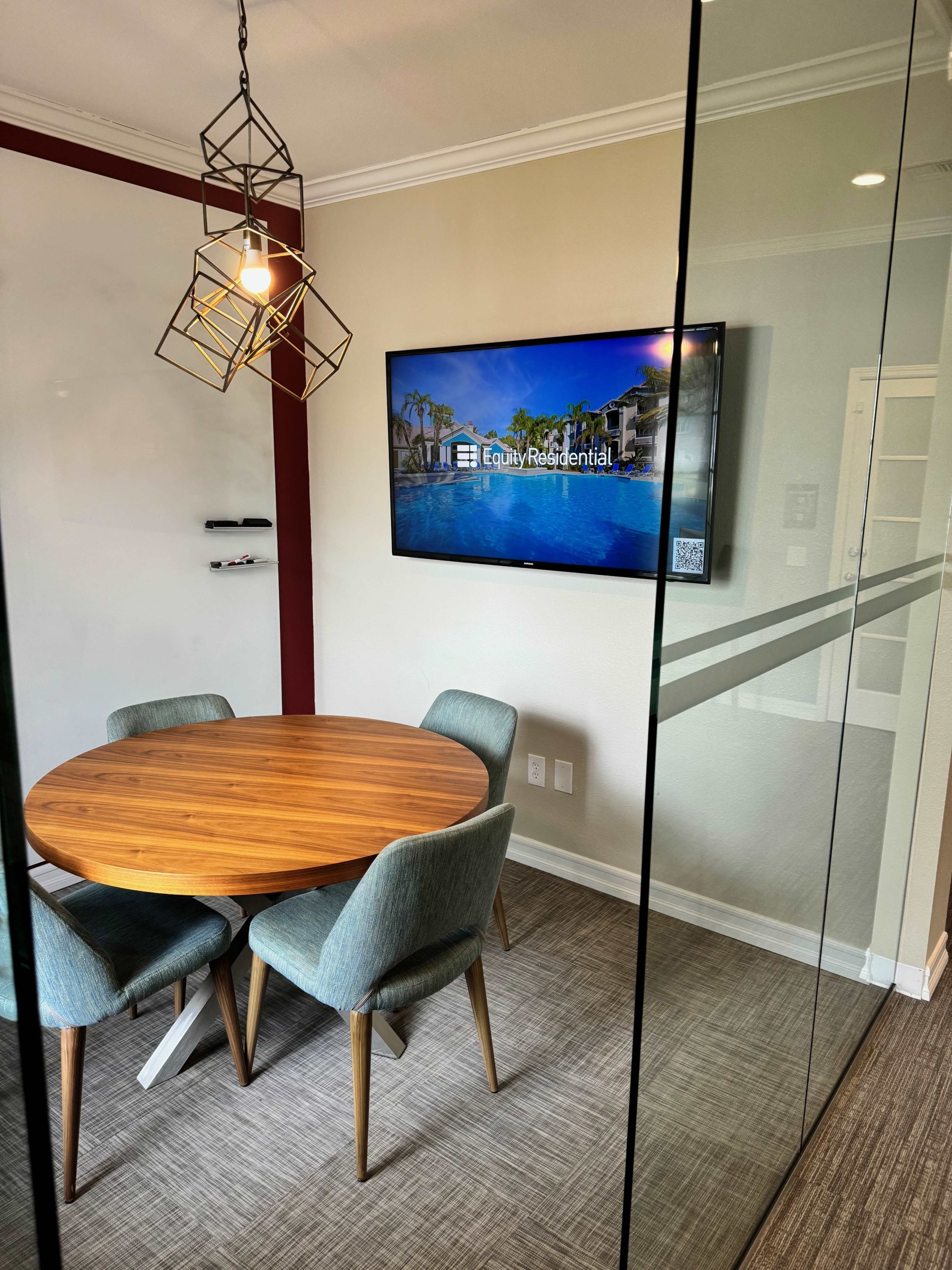 A modern conference room with a round wooden table, four upholstered chairs, and a wall-mounted television displaying a property listing.