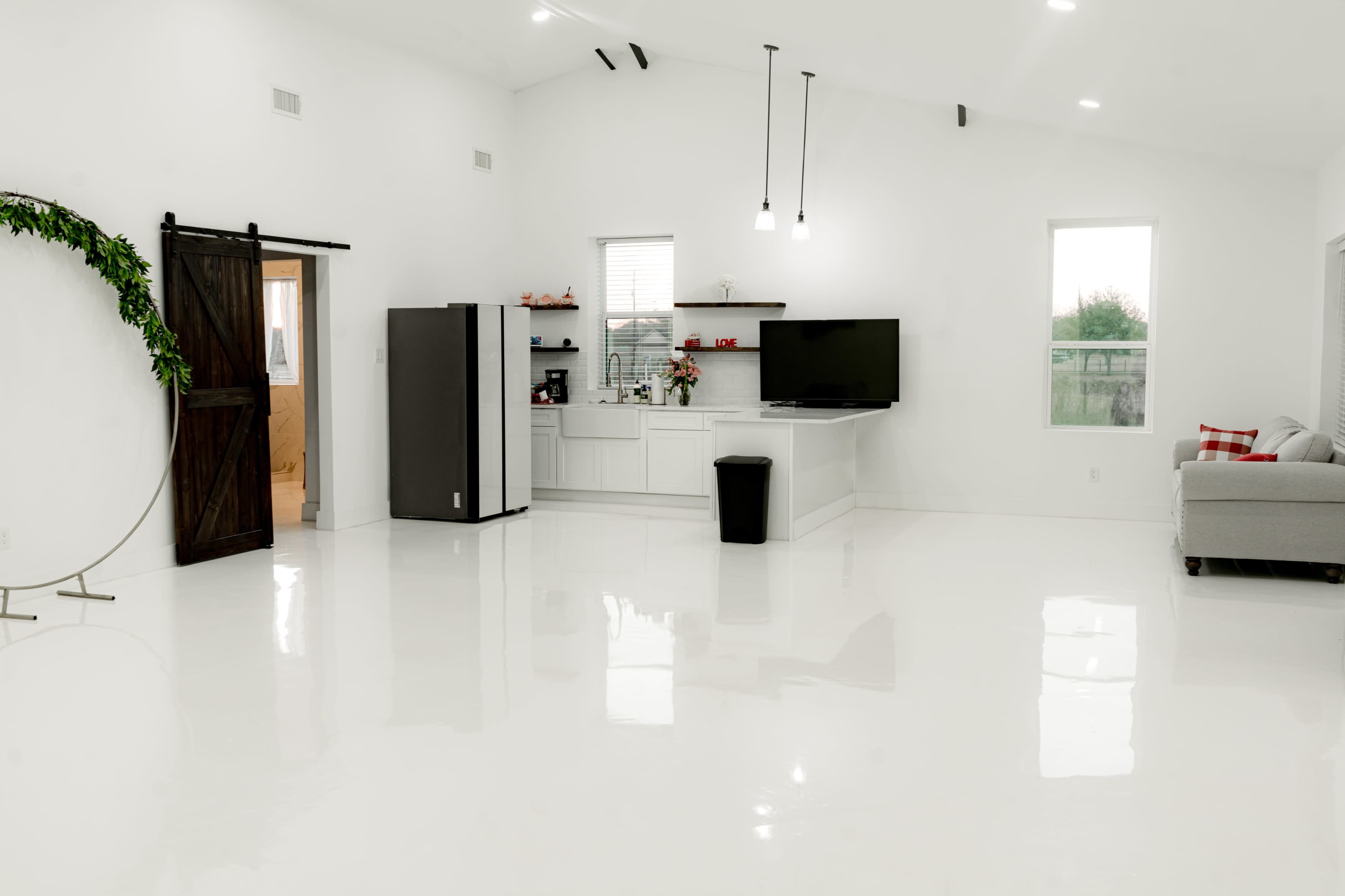The image shows a bright, minimalistic open living space featuring a kitchen area with modern appliances, a television mounted on the wall, and a grey couch.